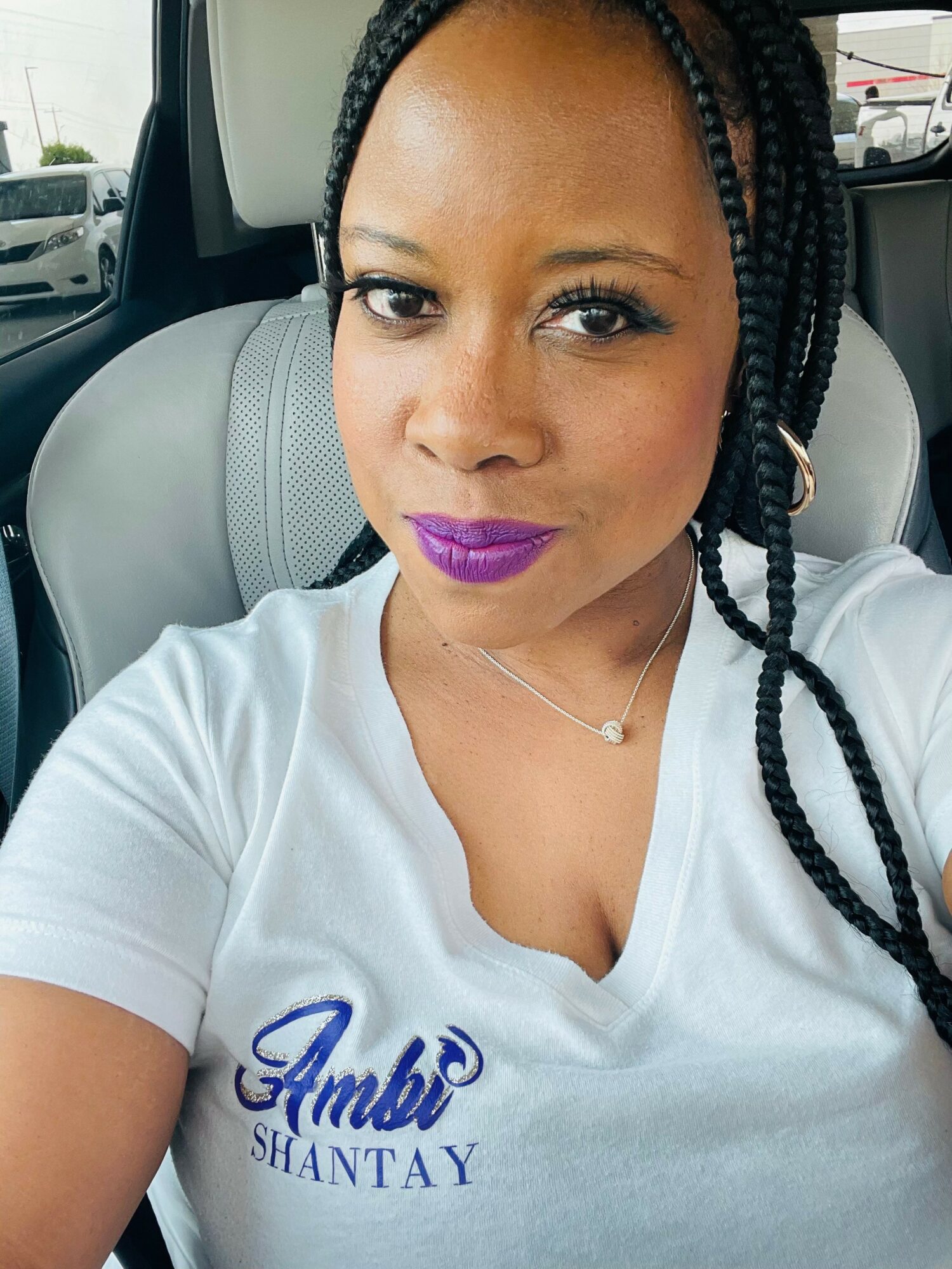 Woman with braided hair and purple lipstick sitting in a car, wearing a white shirt with blue text.