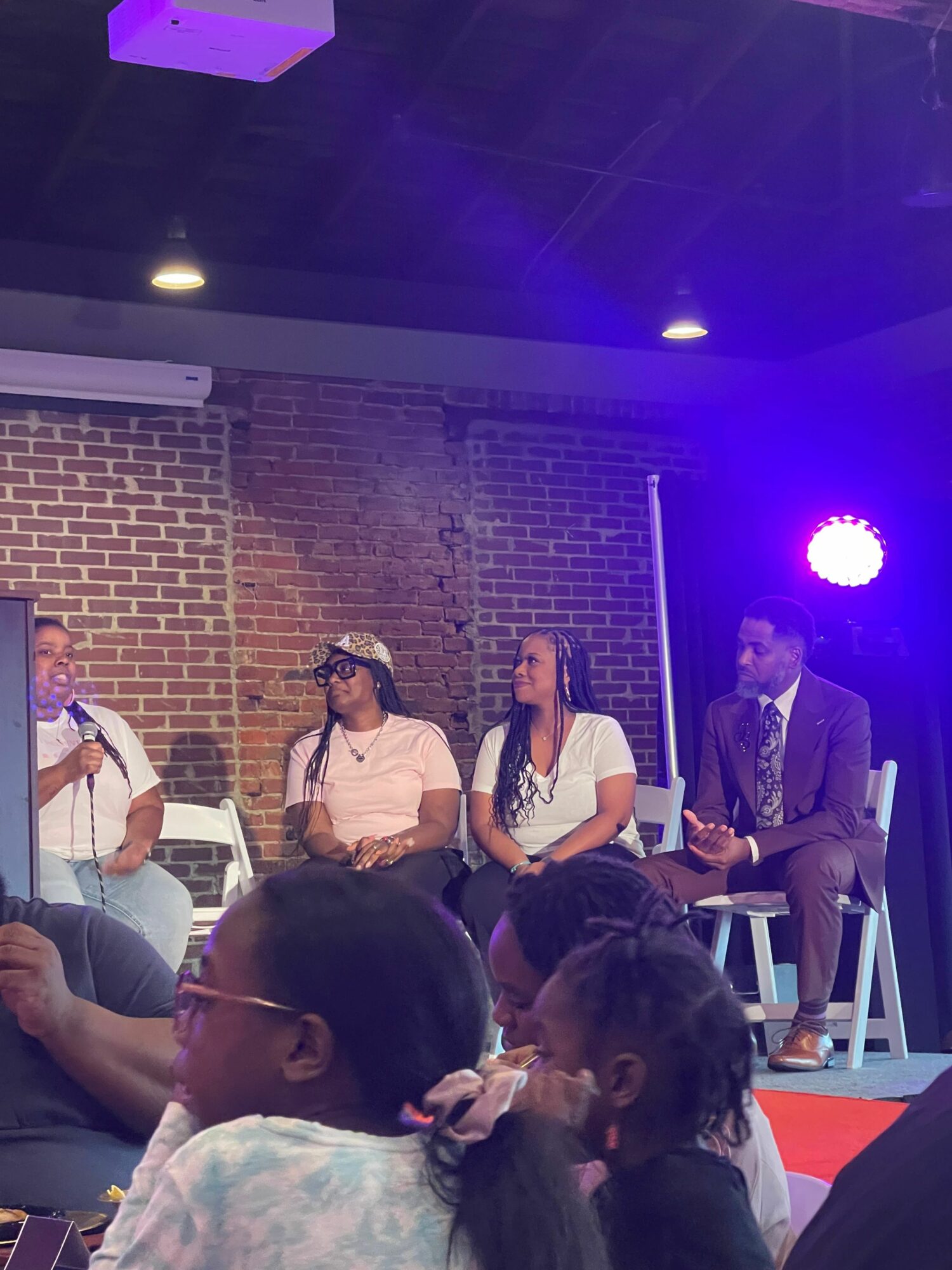 People on stage and in audience, brick wall background, purple stage lighting, some seated and some standing.