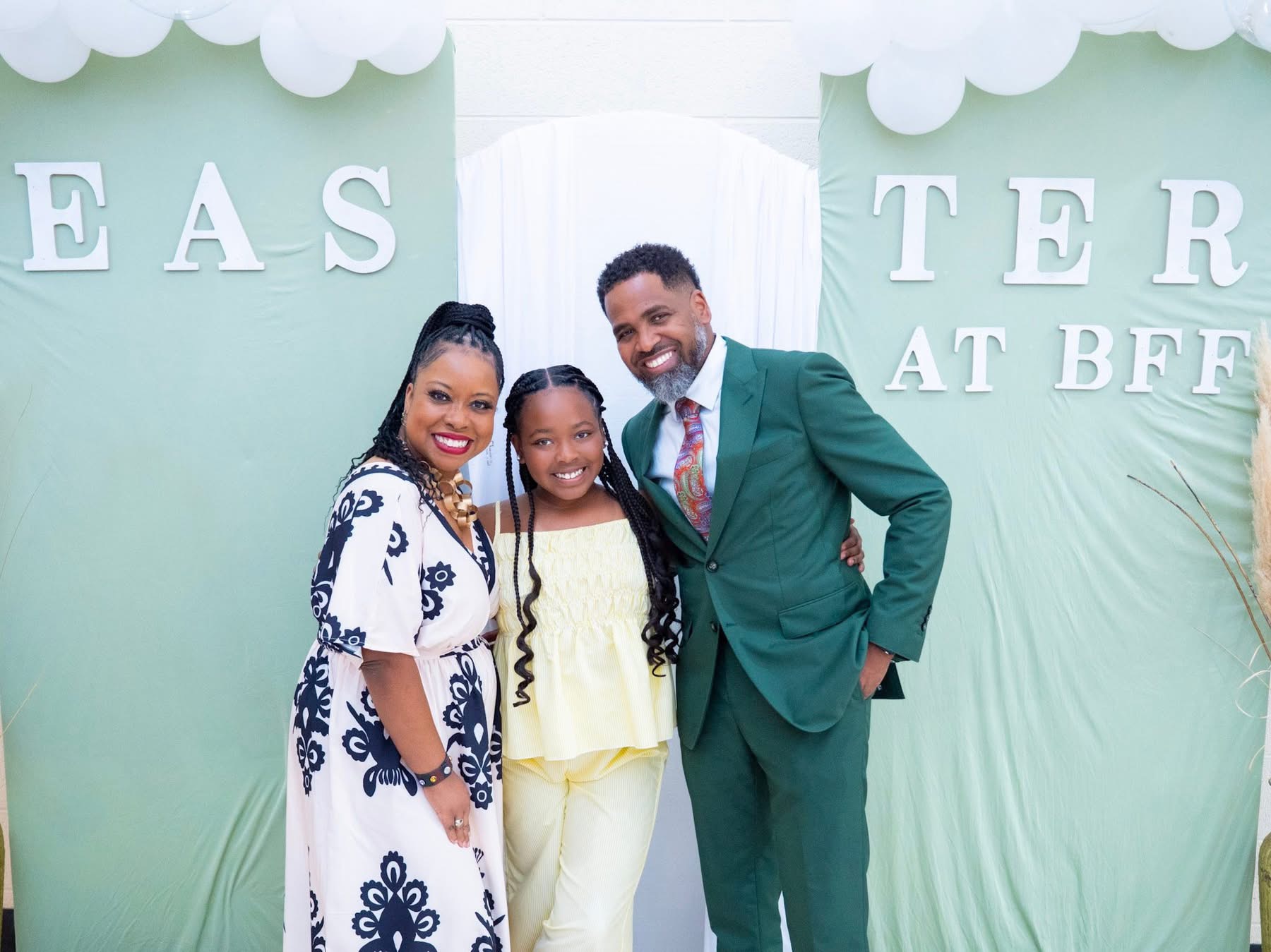Three people smiling at an event, standing in front of a pastel green backdrop with large white letters, one man and two women.