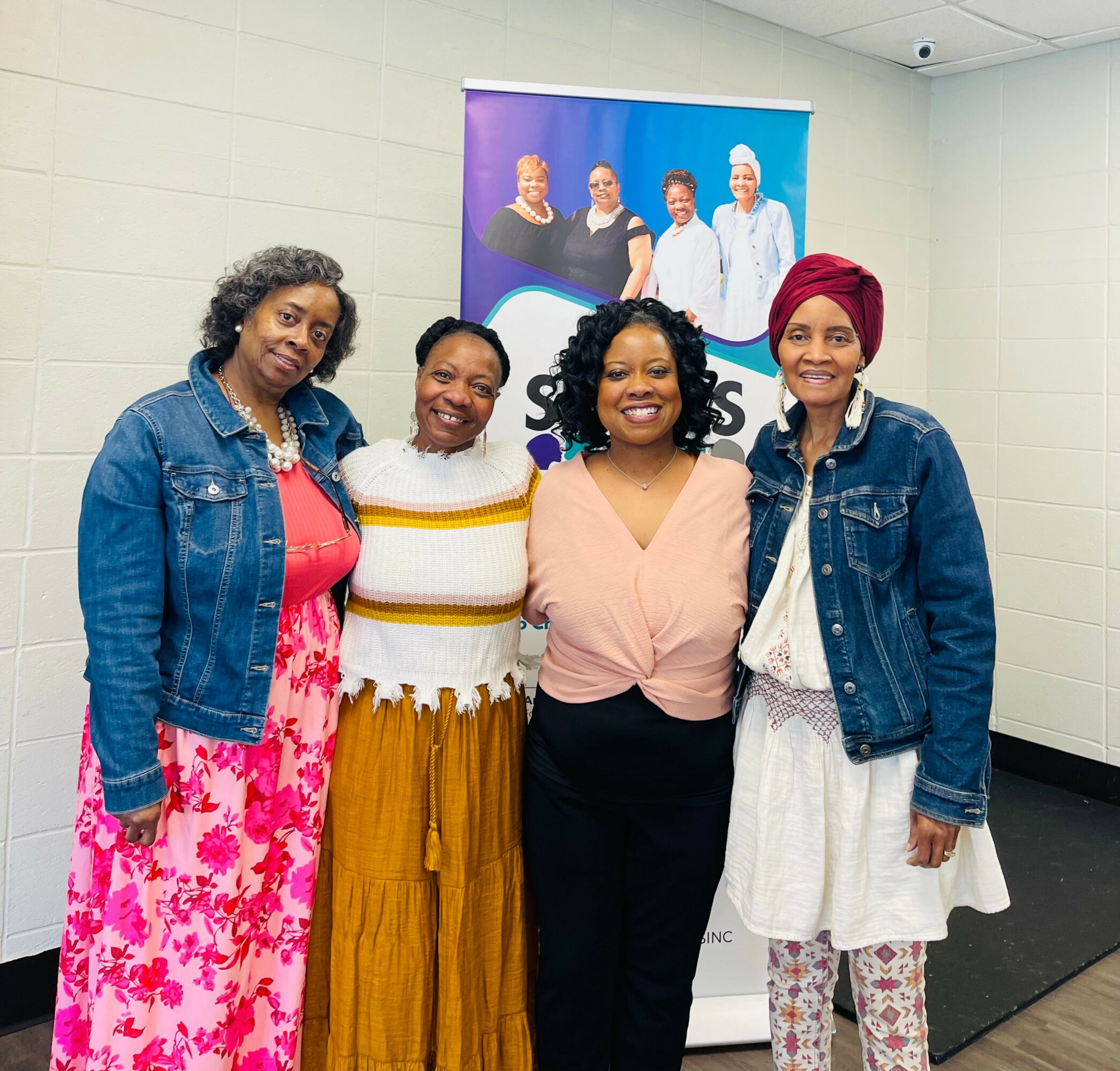 Four women standing together indoors, smiling, with a poster of four people in the background.