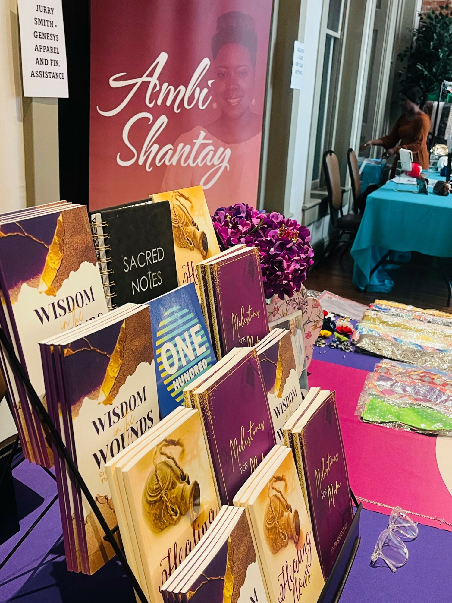 Book display with titles like 'Wisdom,' 'Sacred Notes,' and 'One,' in front of a pink poster with a woman's face and the name 'Ambi Shantay.'