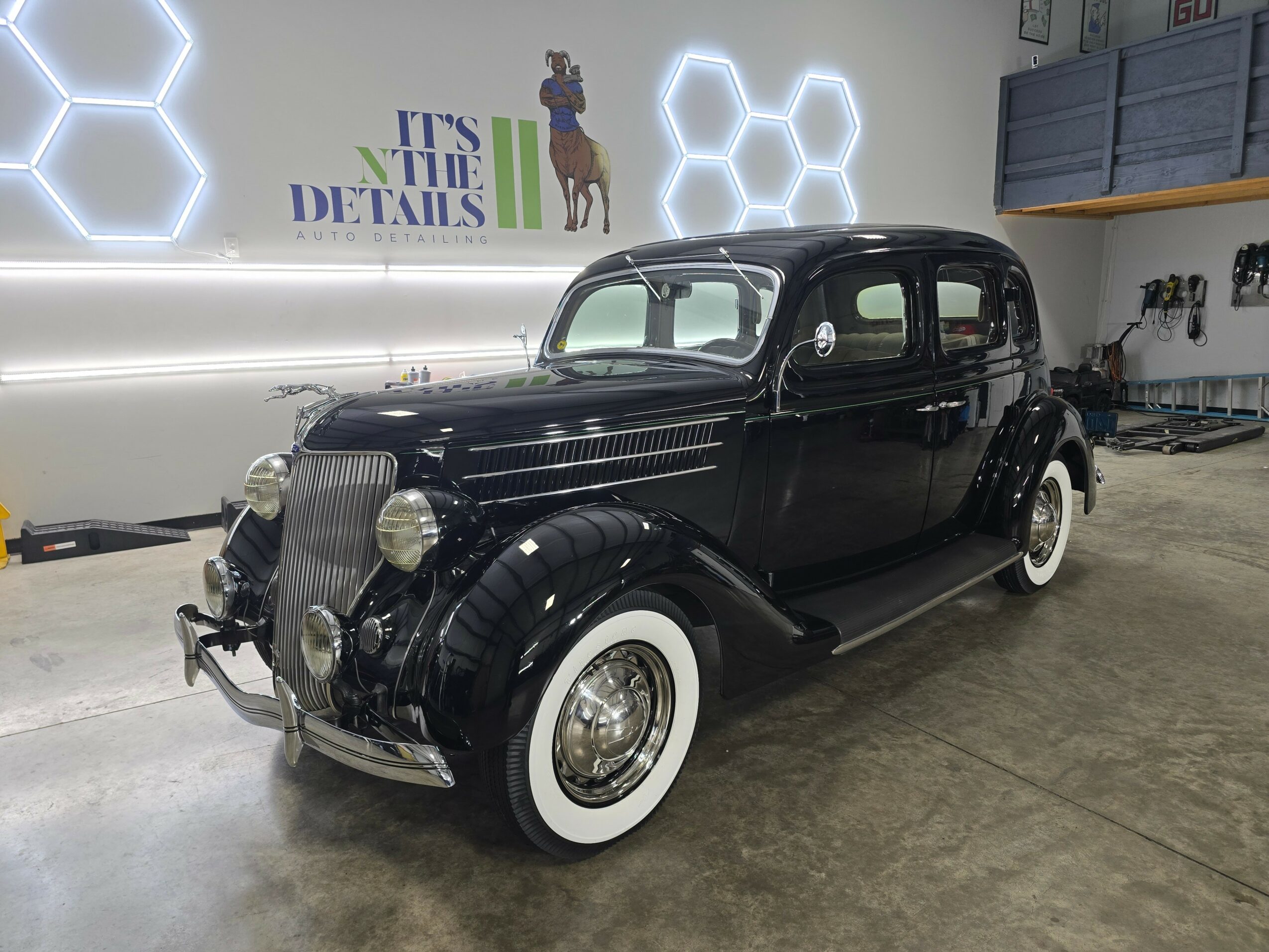 Black vintage car with whitewall tires parked indoors, with hexagonal wall lights and a sign in the background.