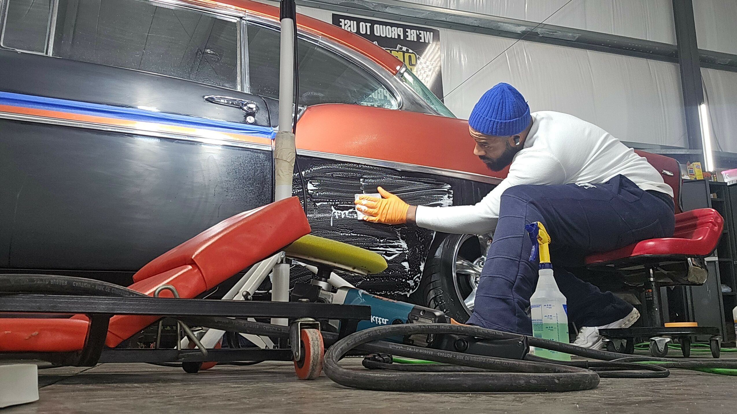 Mechanic working on a car inside a garage, using tools and wearing gloves and a blue cap.