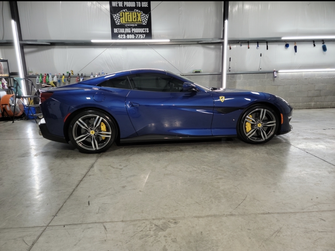 Blue sports car parked indoors, with a sign and equipment in the background, on a concrete floor.