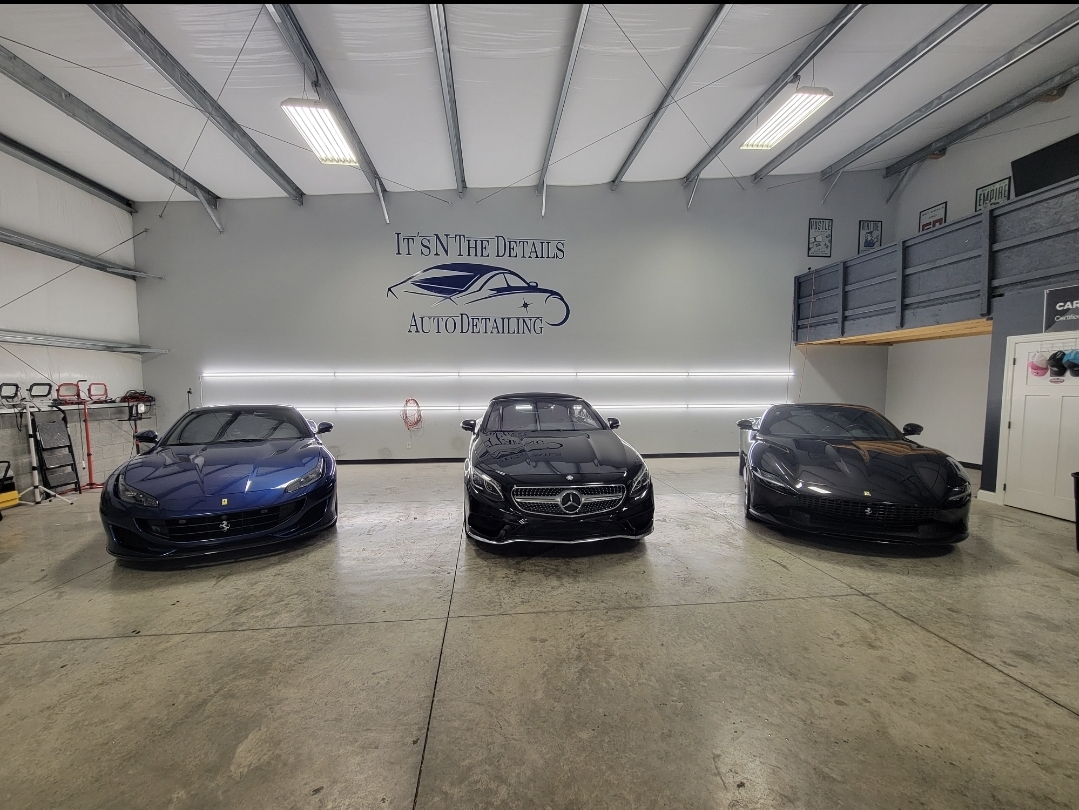 Three luxury cars parked inside a garage with a sign on the wall reading 'It's in the Details Auto Detailing.'