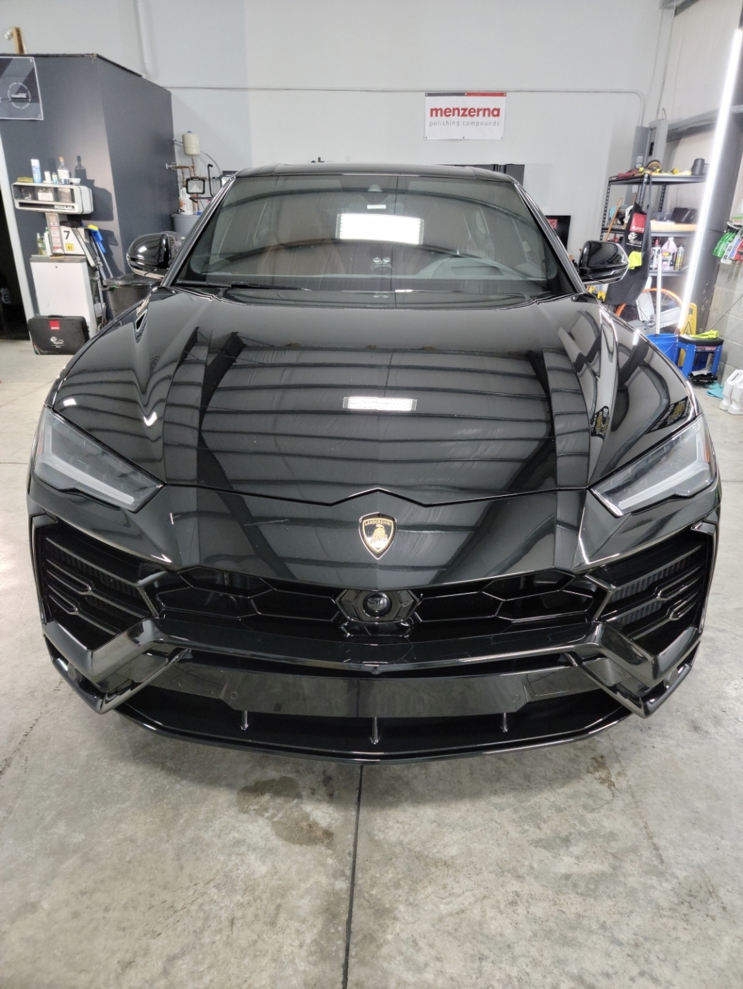Black sports car with sleek design parked inside a garage, reflecting overhead lights, with tools and equipment in the background.
