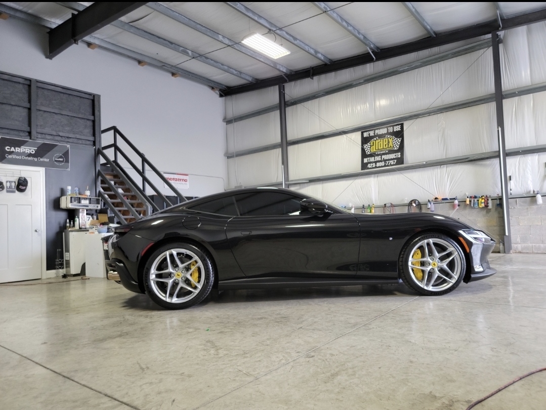Black sports car parked inside a garage with a staircase and tools, industrial setting, high ceiling, metal walls.
