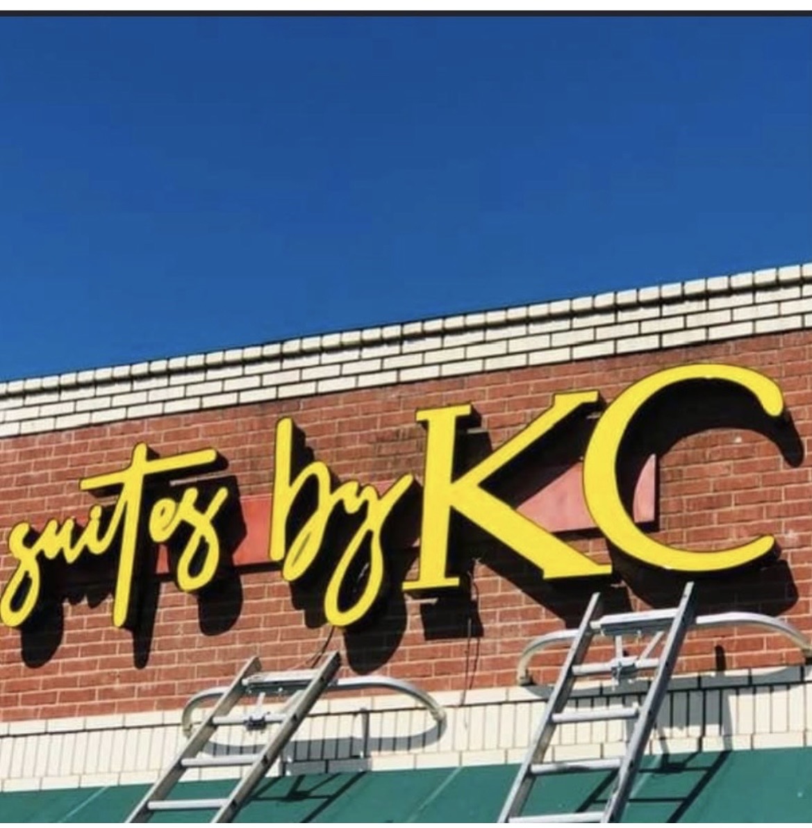 Sign on brick wall with yellow and black text, part of a building exterior, under a blue sky.