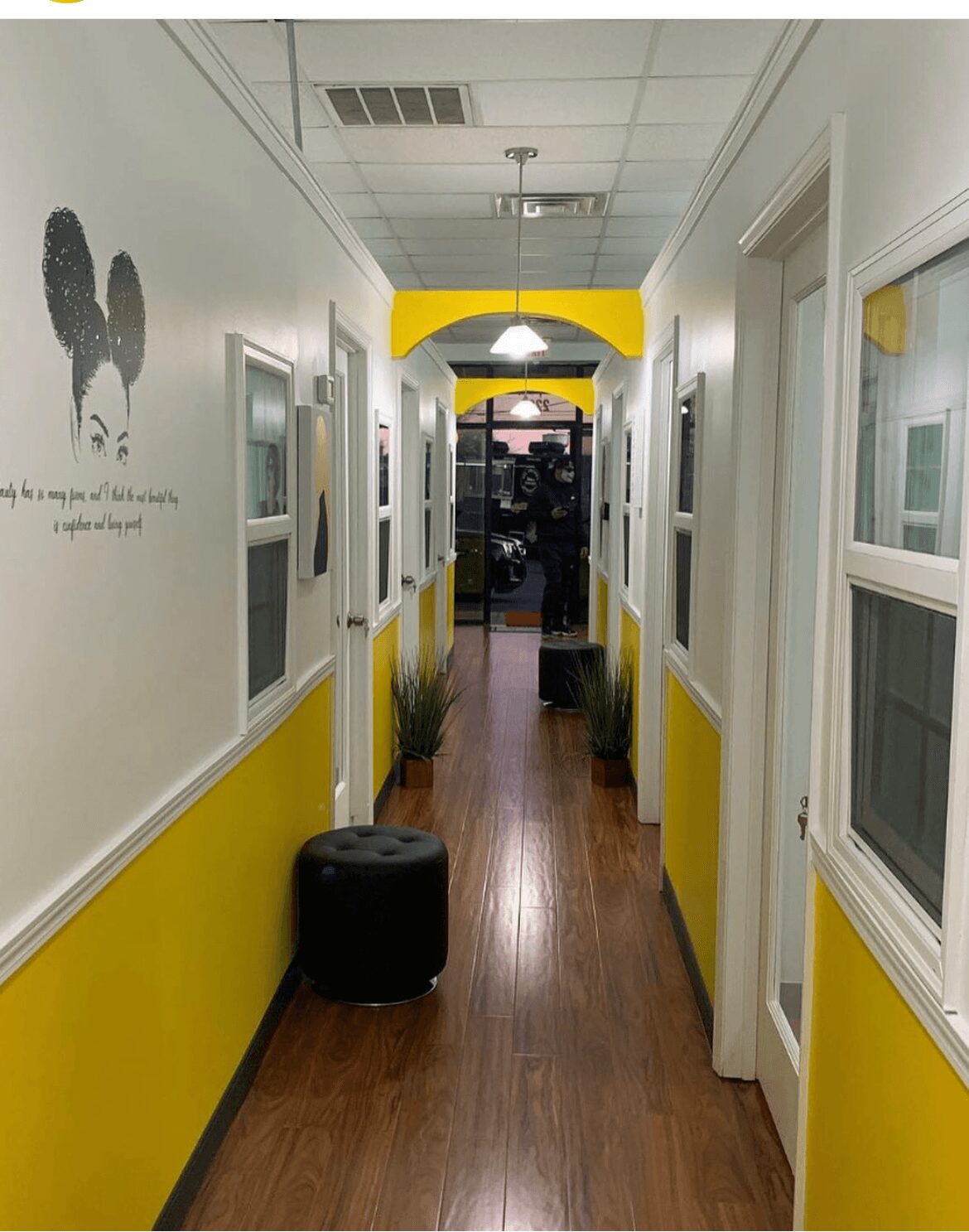 Hallway with yellow and white walls, windows on the right, plants, and a black ottoman, leading to a room with a desk.