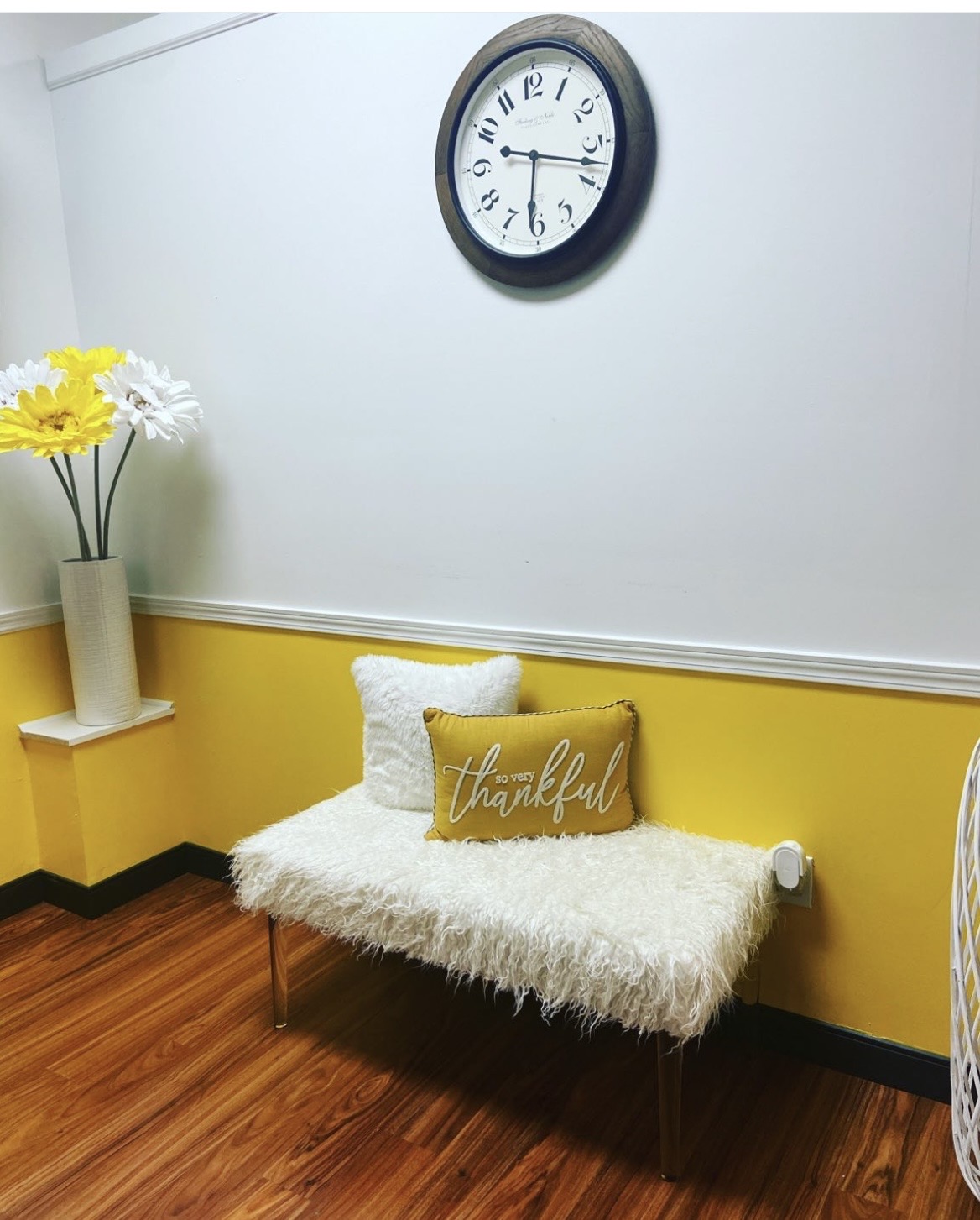 White fluffy bench with a yellow pillow reading 'thankful' in a room with yellow and white walls, a clock, and a vase of flowers.