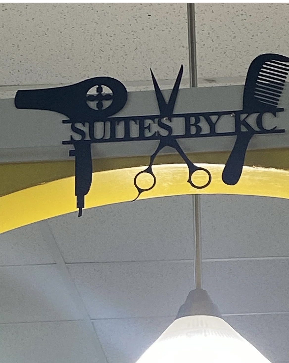 Sign with scissors, comb, and hairdryer silhouette, reading 'SULTES BY KC', hanging from ceiling with a light fixture below.