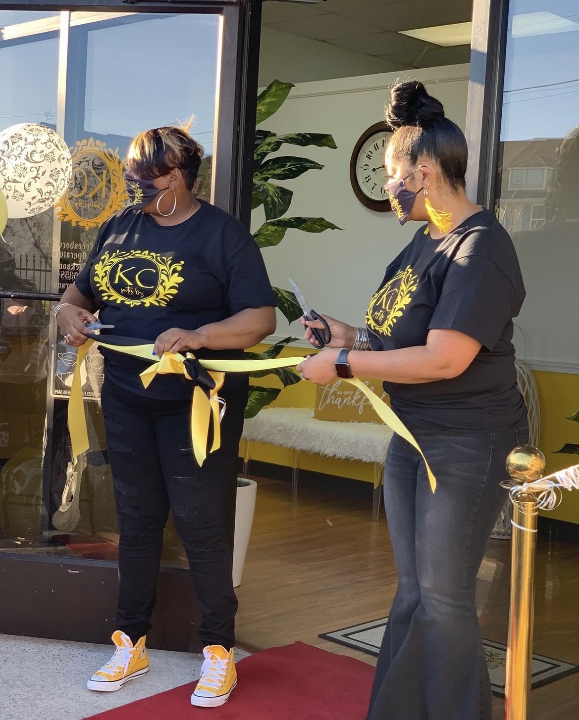 Two women in black shirts with yellow logos cut a yellow ribbon outside a building, holding scissors and a ribbon. Bright sunlight.