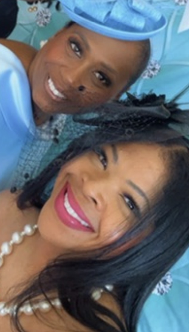 Two women smiling, one wearing a blue hat and dress, the other with black hair and a pearl necklace.