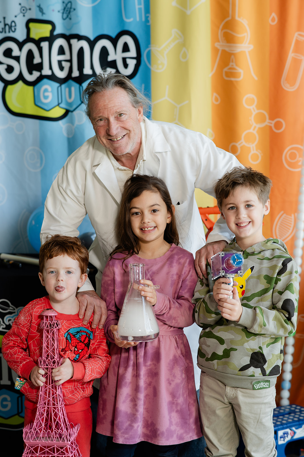 Smiling man with three children holding science-related objects in front of a colorful science-themed backdrop.