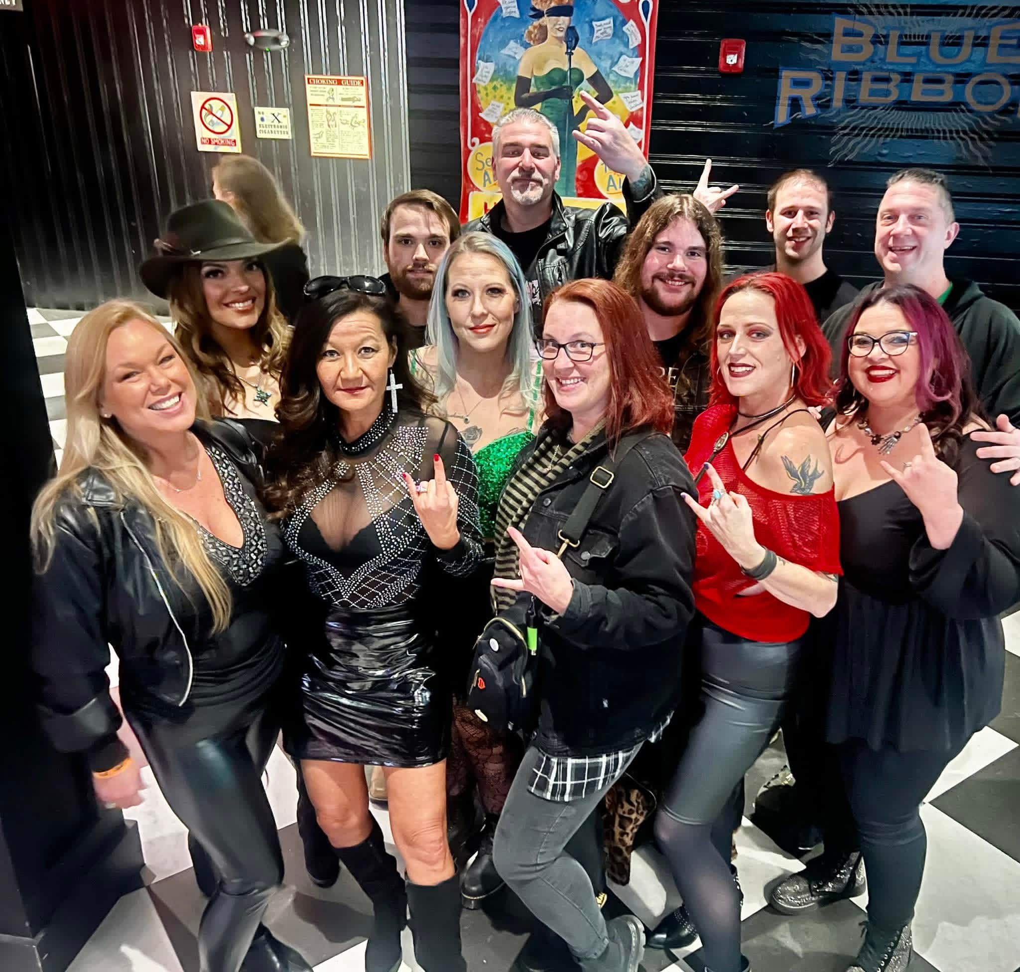 Group of people smiling and posing together indoors, some making hand gestures, with a colorful background and a bus. Word 'RIBBO' visible.