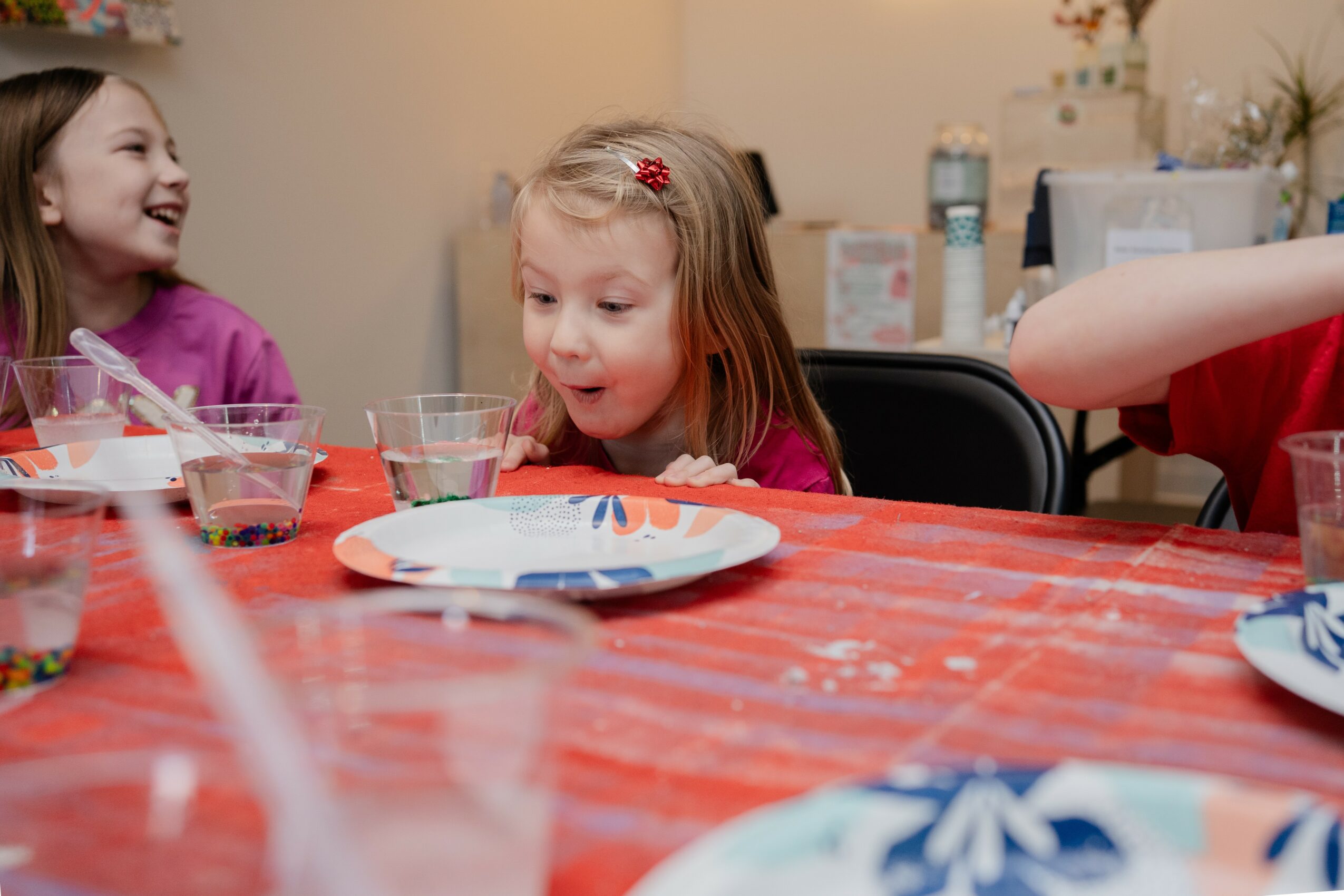 Two young girls sitting at a table with party plates and drinks, smiling and laughing, celebrating a birthday or special occasion.