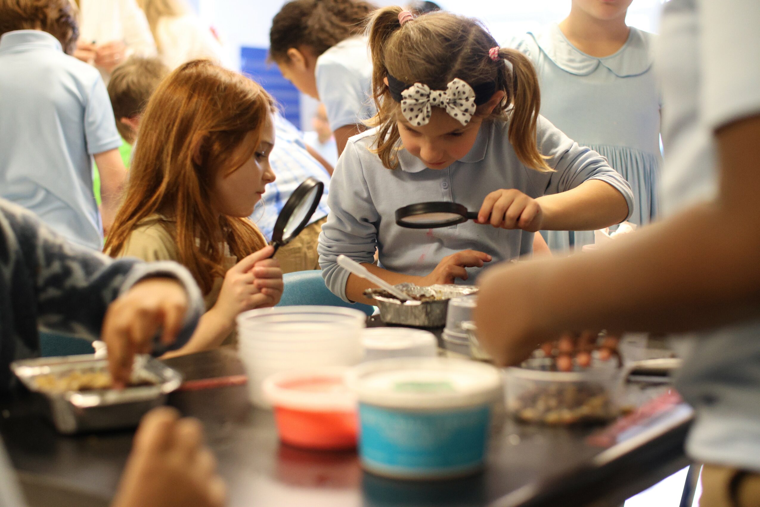 Children gathered around a table, examining objects with magnifying glasses, in a classroom or activity setting.