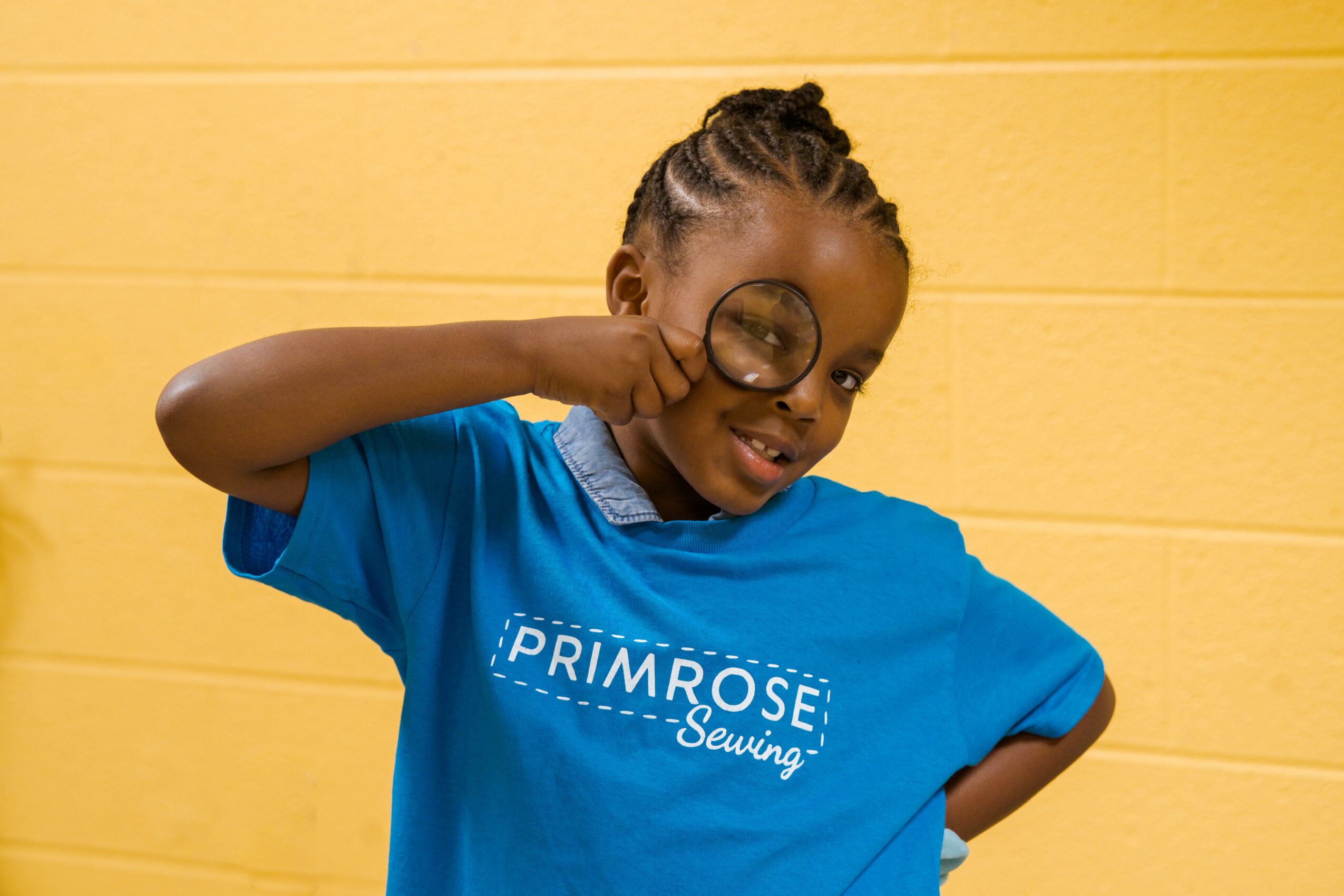 Child with braided hair wearing a blue shirt and large glasses, smiling and posing with a fist near face, yellow background.