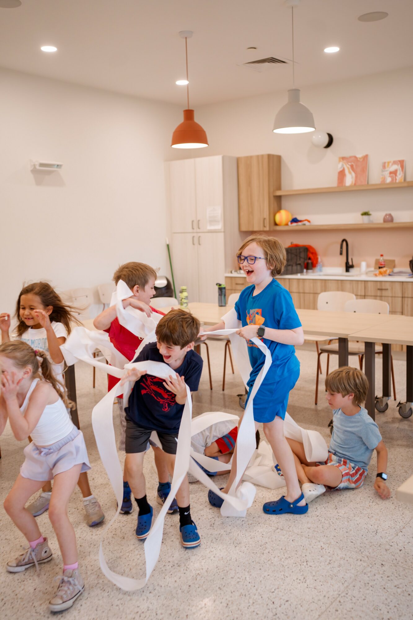 Children playing with a long white ribbon in a bright, modern kitchen and dining area with white walls and wooden cabinets.