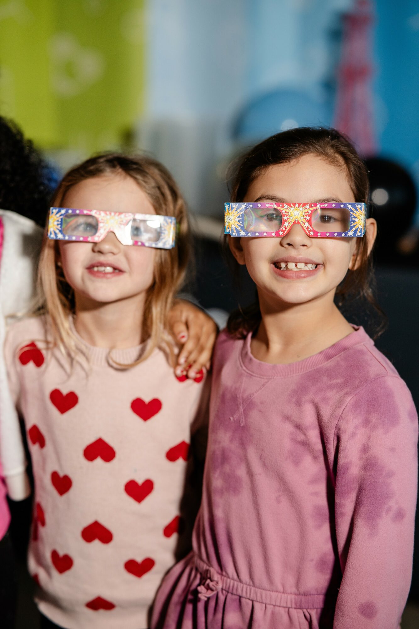 Two children wearing colorful glasses and smiling, standing close together in a colorful indoor setting.