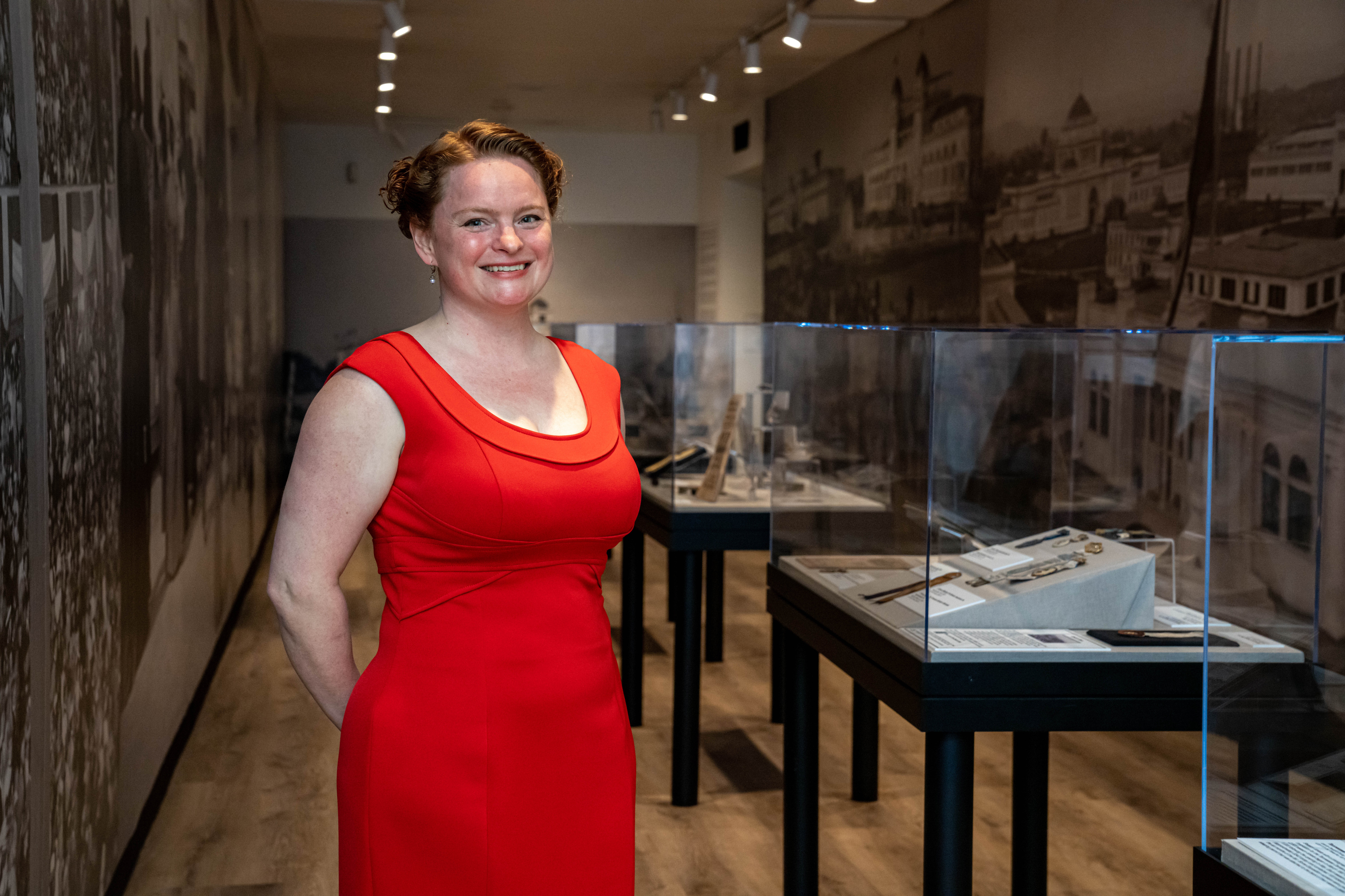 Woman in red dress smiling in museum exhibit with display cases and historical photos, wooden floor, and dim lighting.