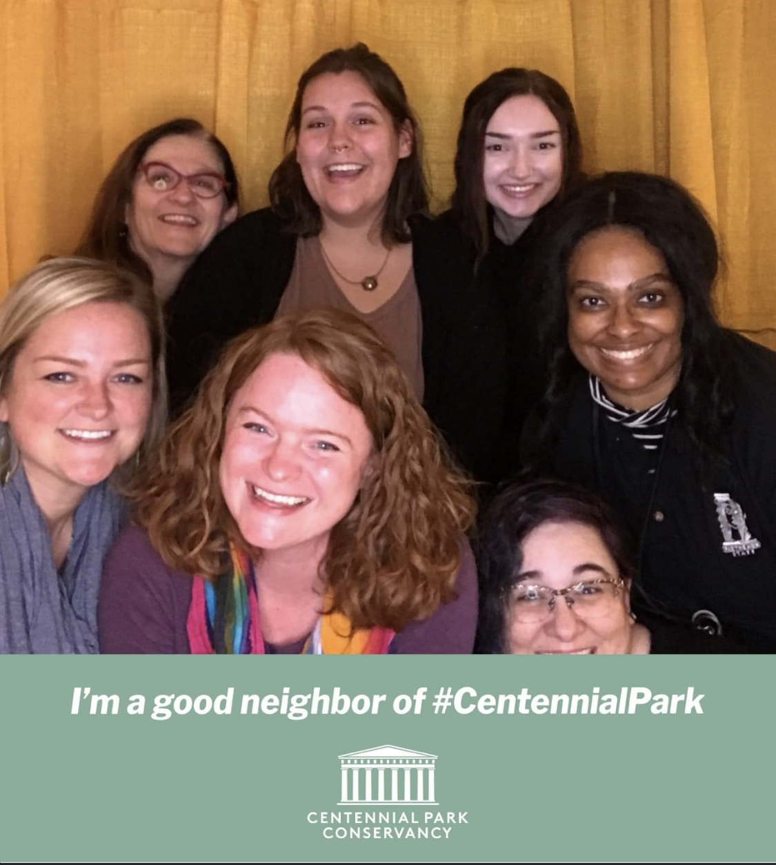 Group of seven women smiling in front of a yellow curtain, with a green banner below reading 'I'm a good neighbor of #CentennialPark' and a logo.