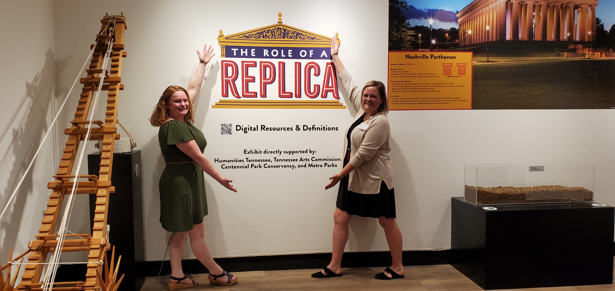 Two women stand in front of a wall display with a sign about replicas, with a model of a building on the right and a poster of a monument on the wall. The women are smiling and pointing at the display.