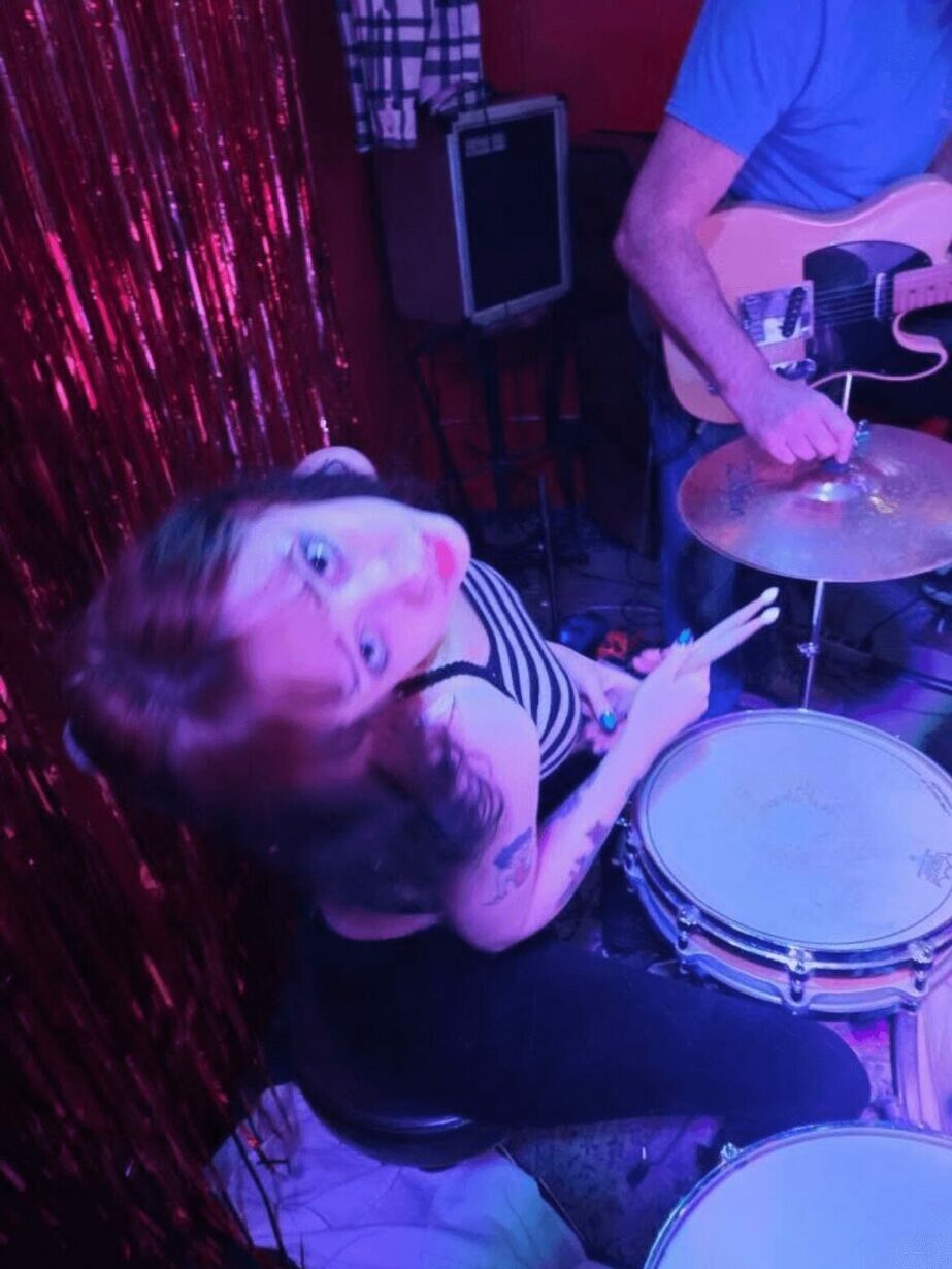 Child playing drums with a person playing guitar in a colorful setting, red and purple background, child holding drumsticks.