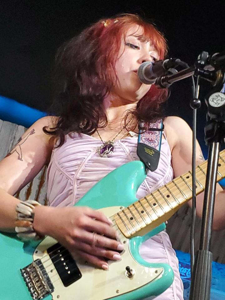 Female musician with red and black hair singing into a microphone while playing a turquoise electric guitar.