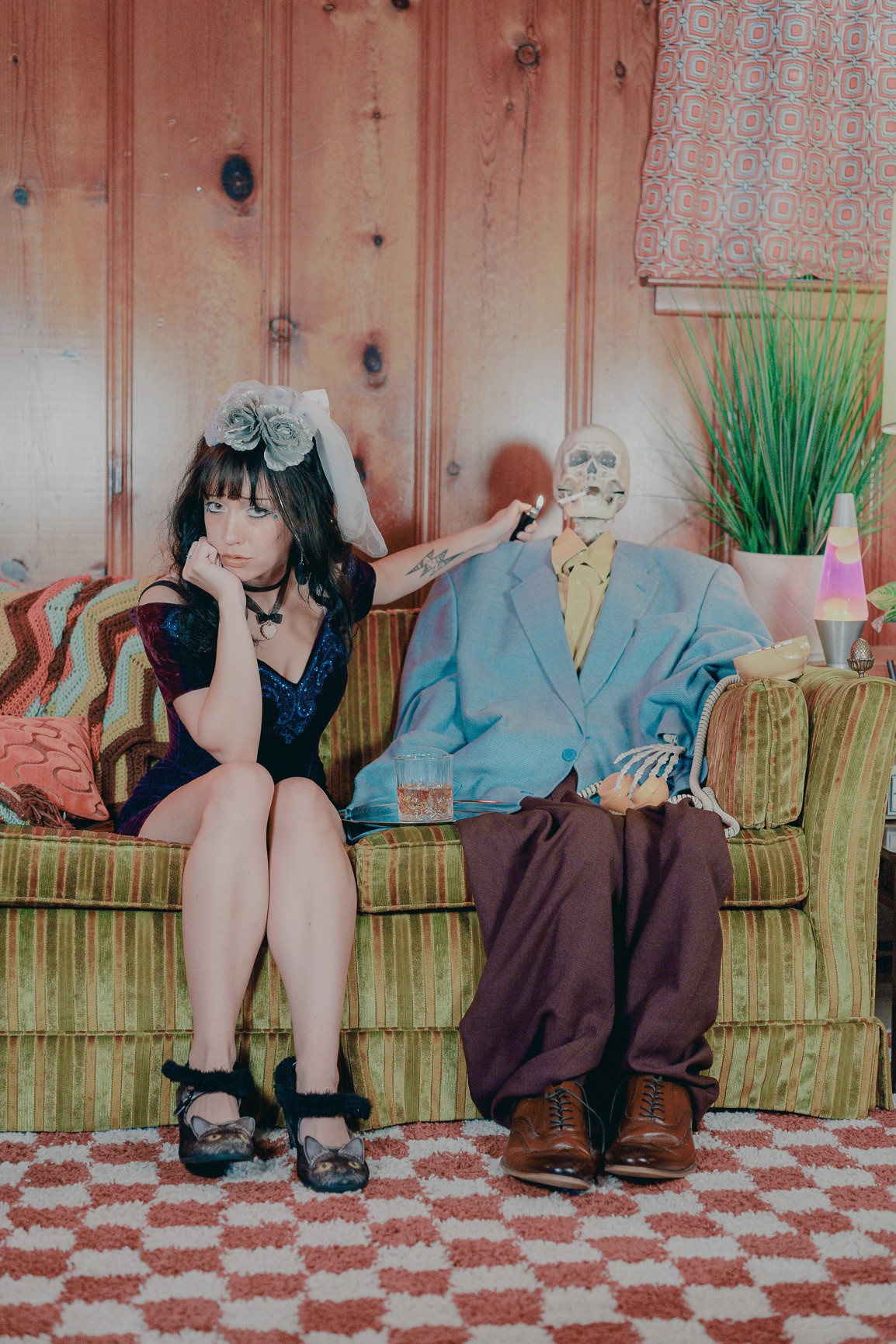 Girl with a large bow in her hair sitting on a sofa next to a skeleton dressed in a suit, in a room with wooden walls.