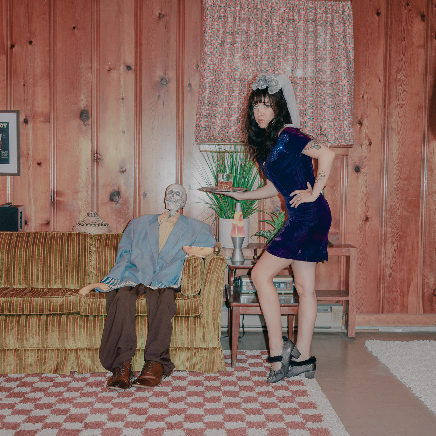 A woman in a blue dress and headband poses next to a mannequin with a paper bag face in a wooden room.