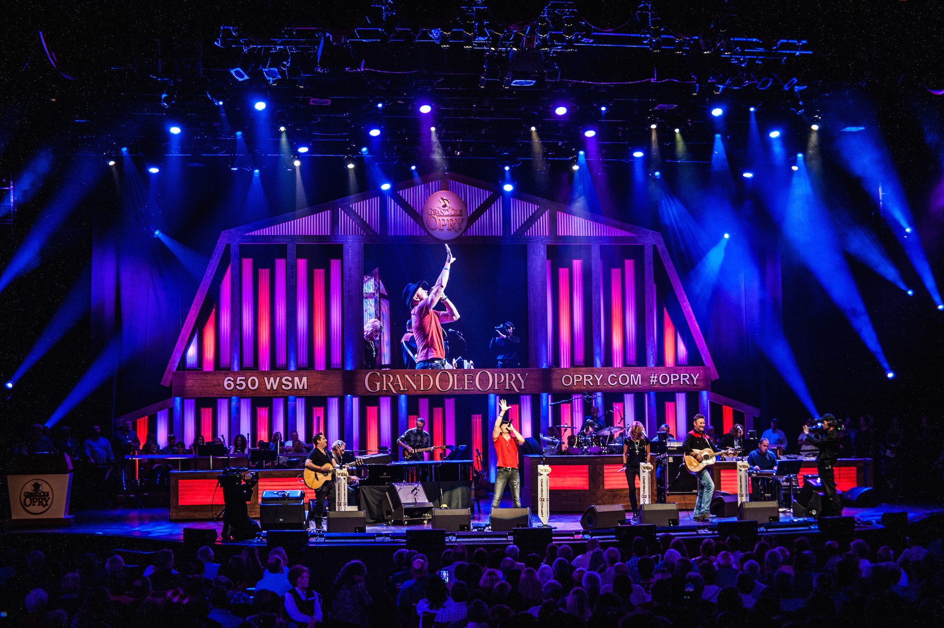Stage with performers, bright lights, and large screens displaying text and images, audience watching the event.