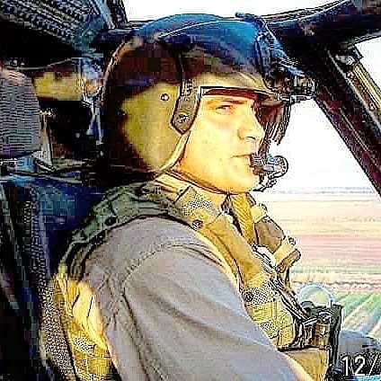 Person in pilot uniform and helmet sitting in aircraft cockpit, looking to the side, with landscape visible outside.