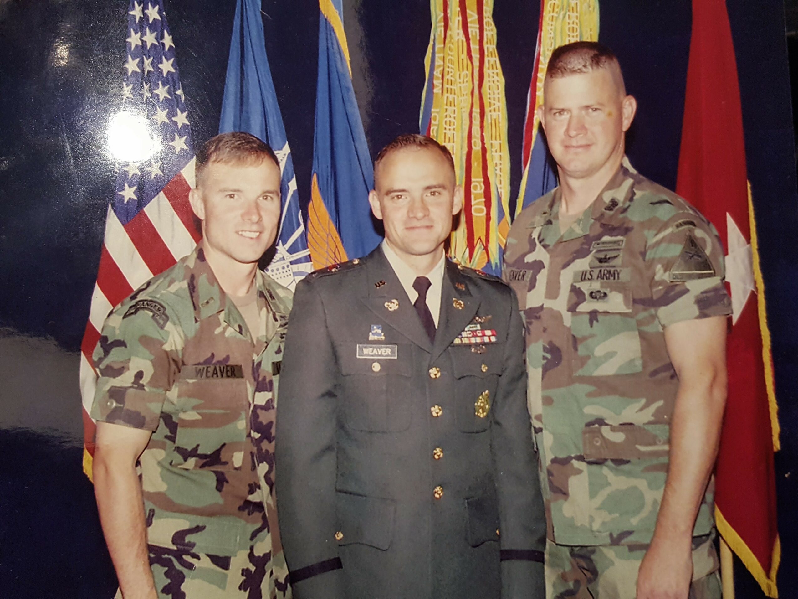 Three military personnel in uniform standing in front of flags, smiling for a photo.