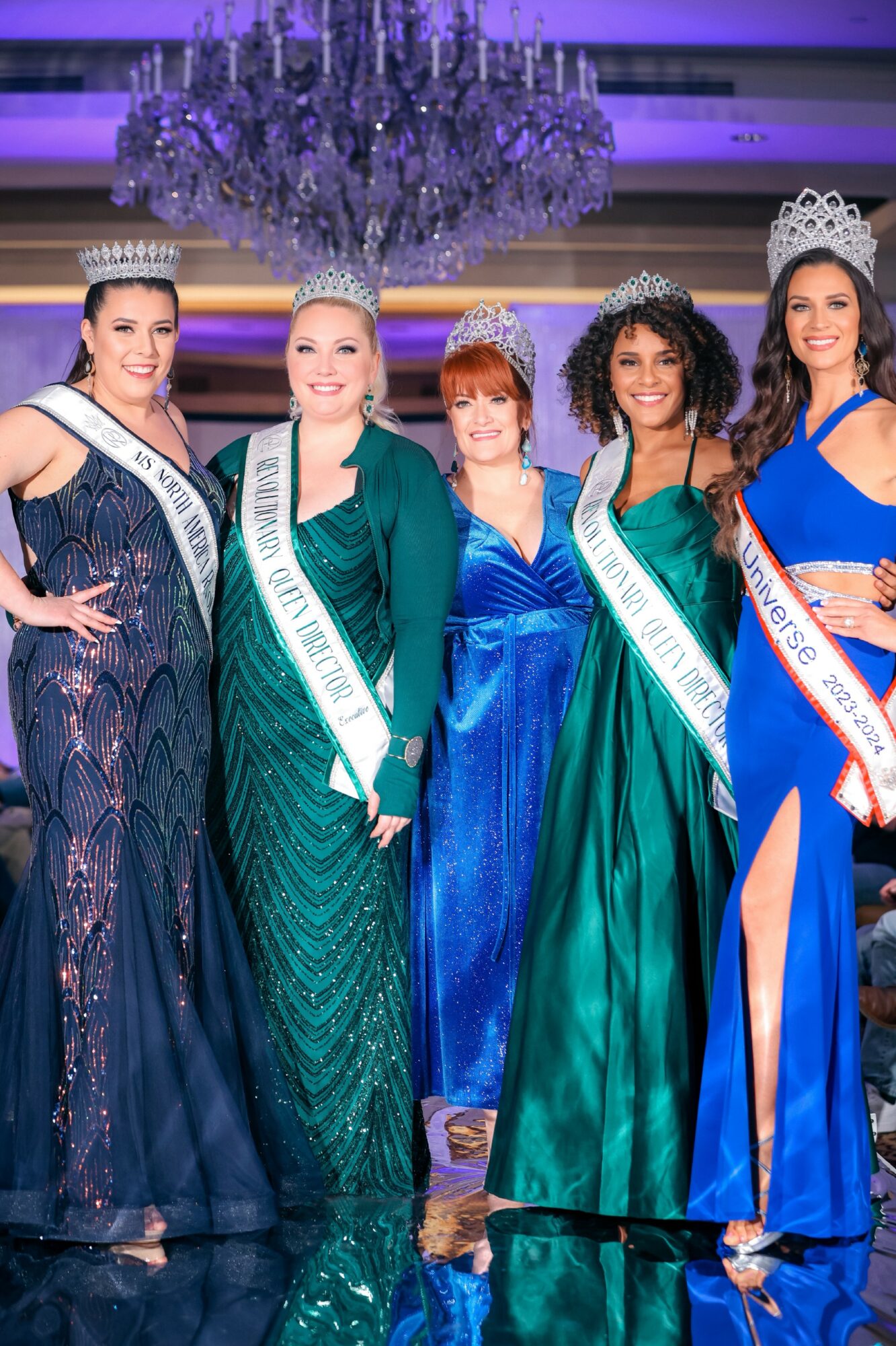Five women in evening gowns and tiaras, wearing sashes, standing in a decorated room with a chandelier.