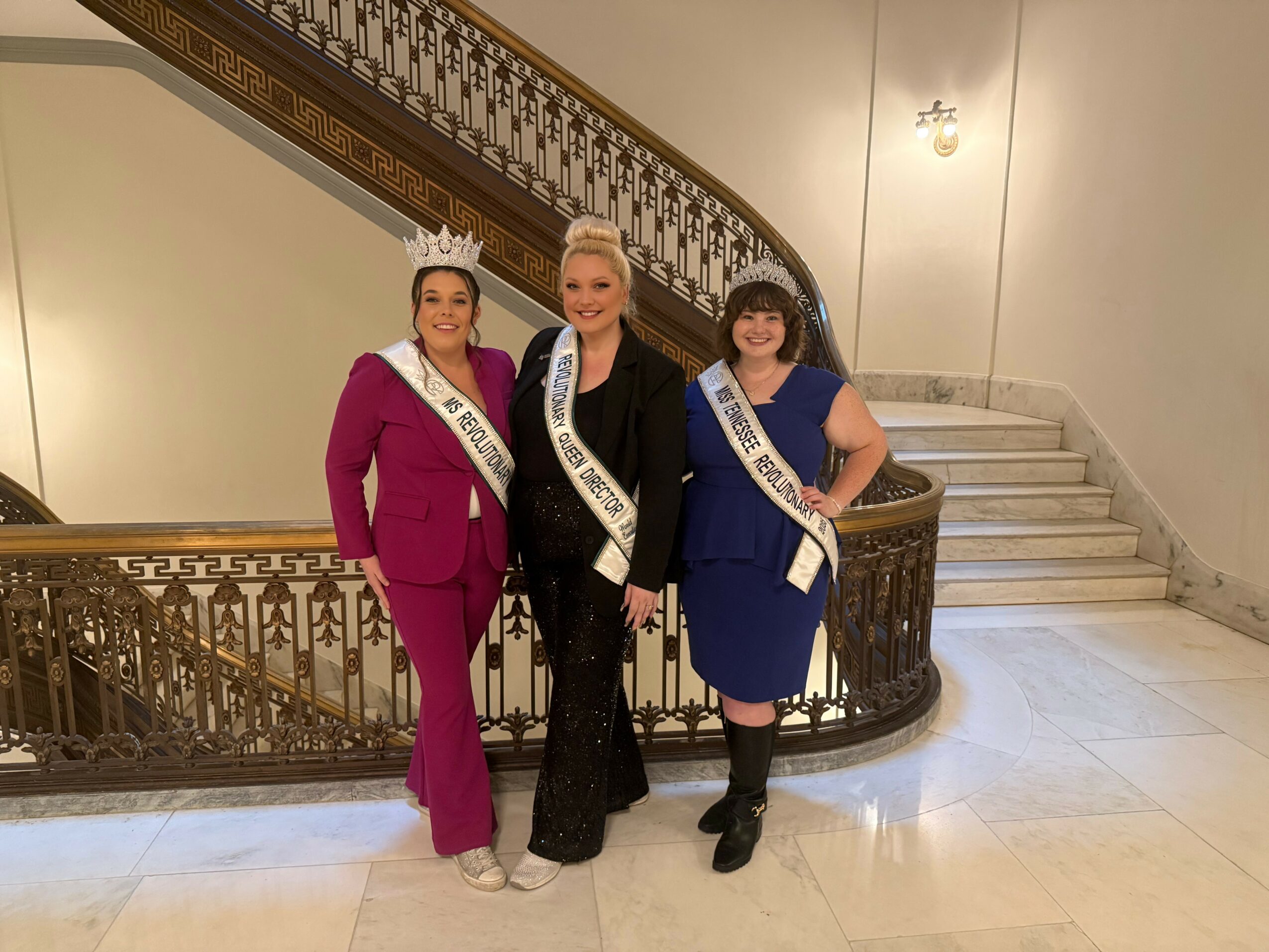 Three women wearing sashes and crowns standing on a staircase railing, smiling at the camera.