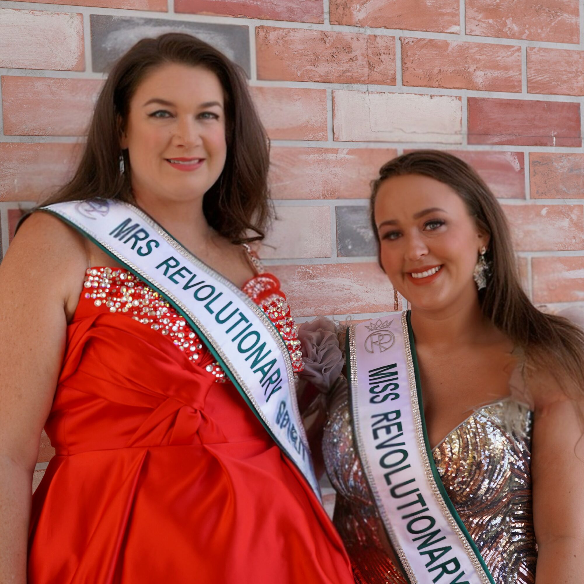 Two women wearing sashes that read 'Mrs. Revolutionary' and 'Mrs. Revolution' stand against a brick wall, smiling.