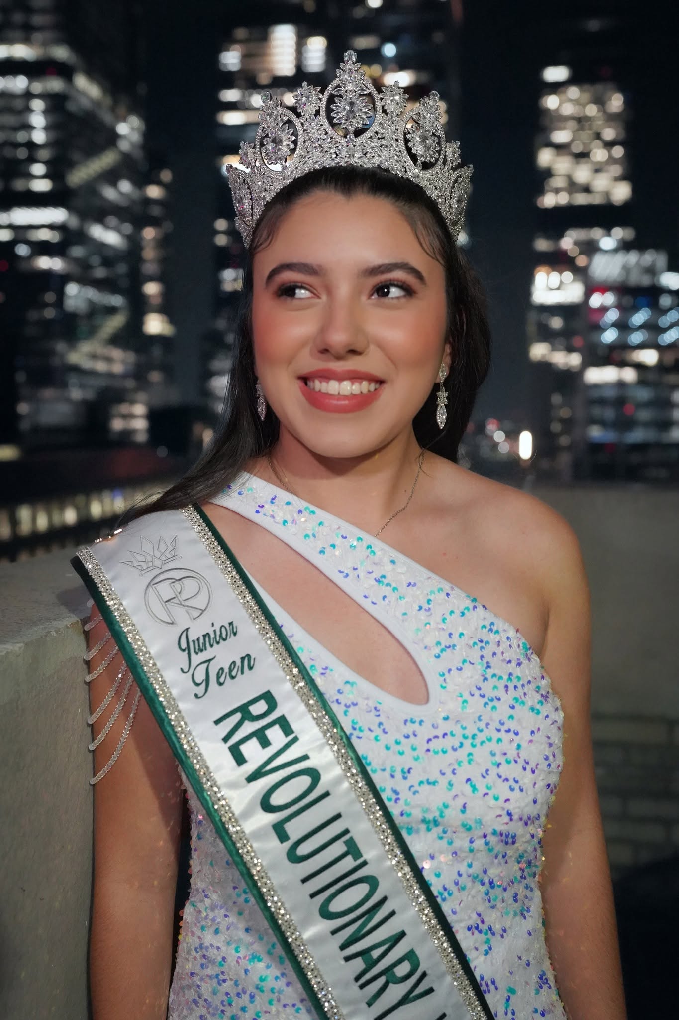 Young woman wearing a crown and sash, smiling, cityscape background at night, wearing a white dress with colorful dots.