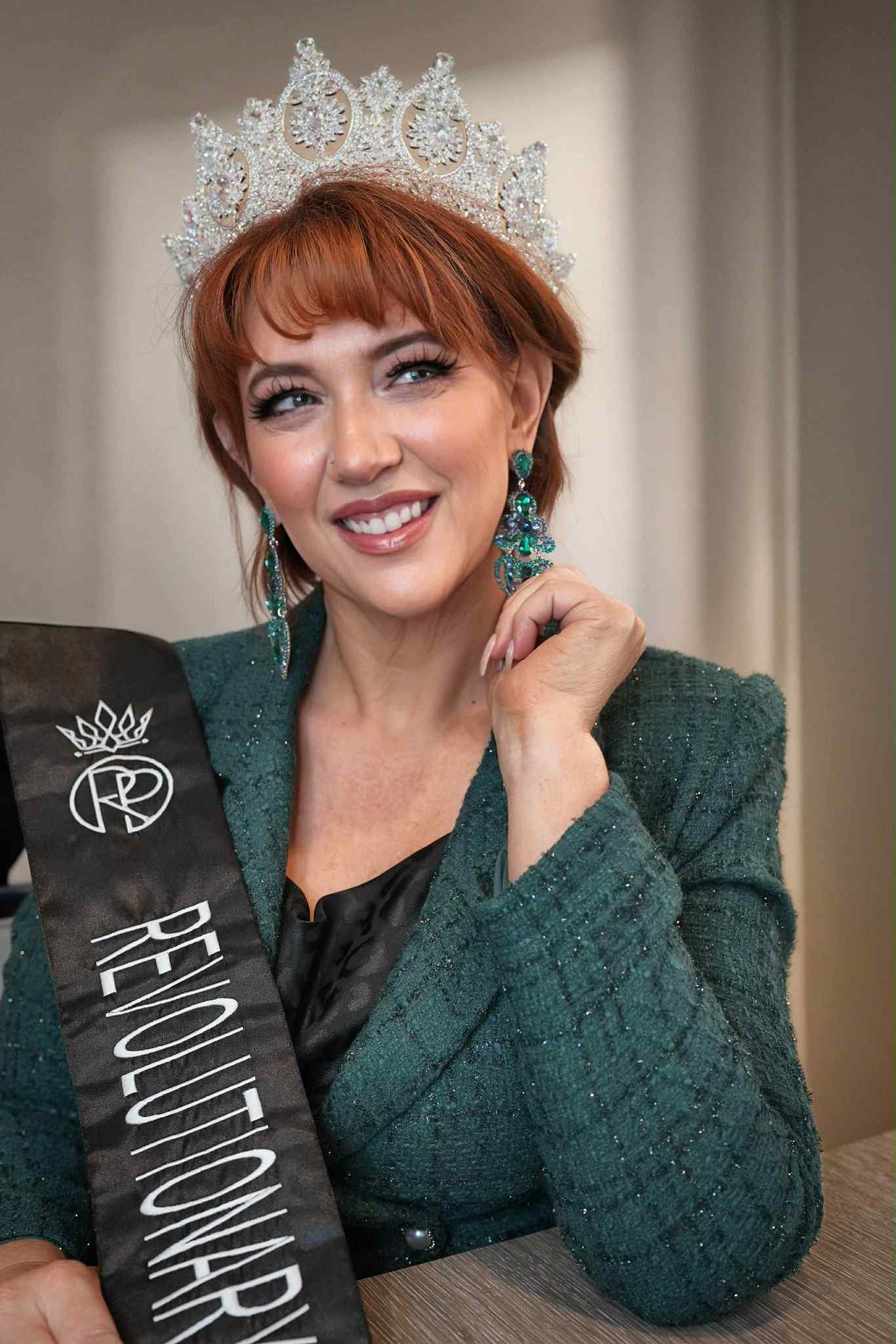 Woman with red hair wearing a crown and sash, smiling, in formal attire, indoors.