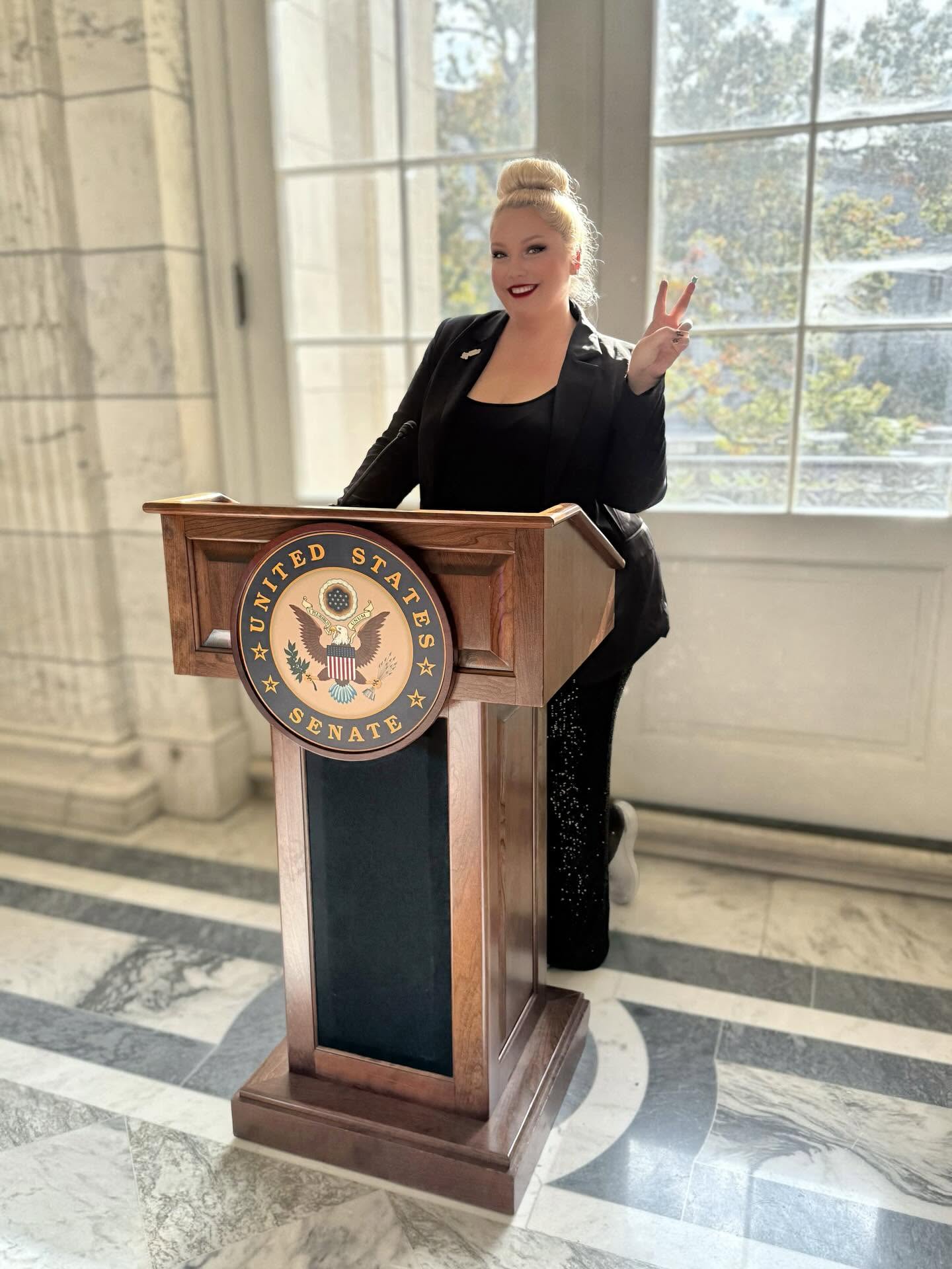 Woman standing behind a podium with a U.S. Senate emblem, smiling and making a peace sign, in a bright room with large windows.