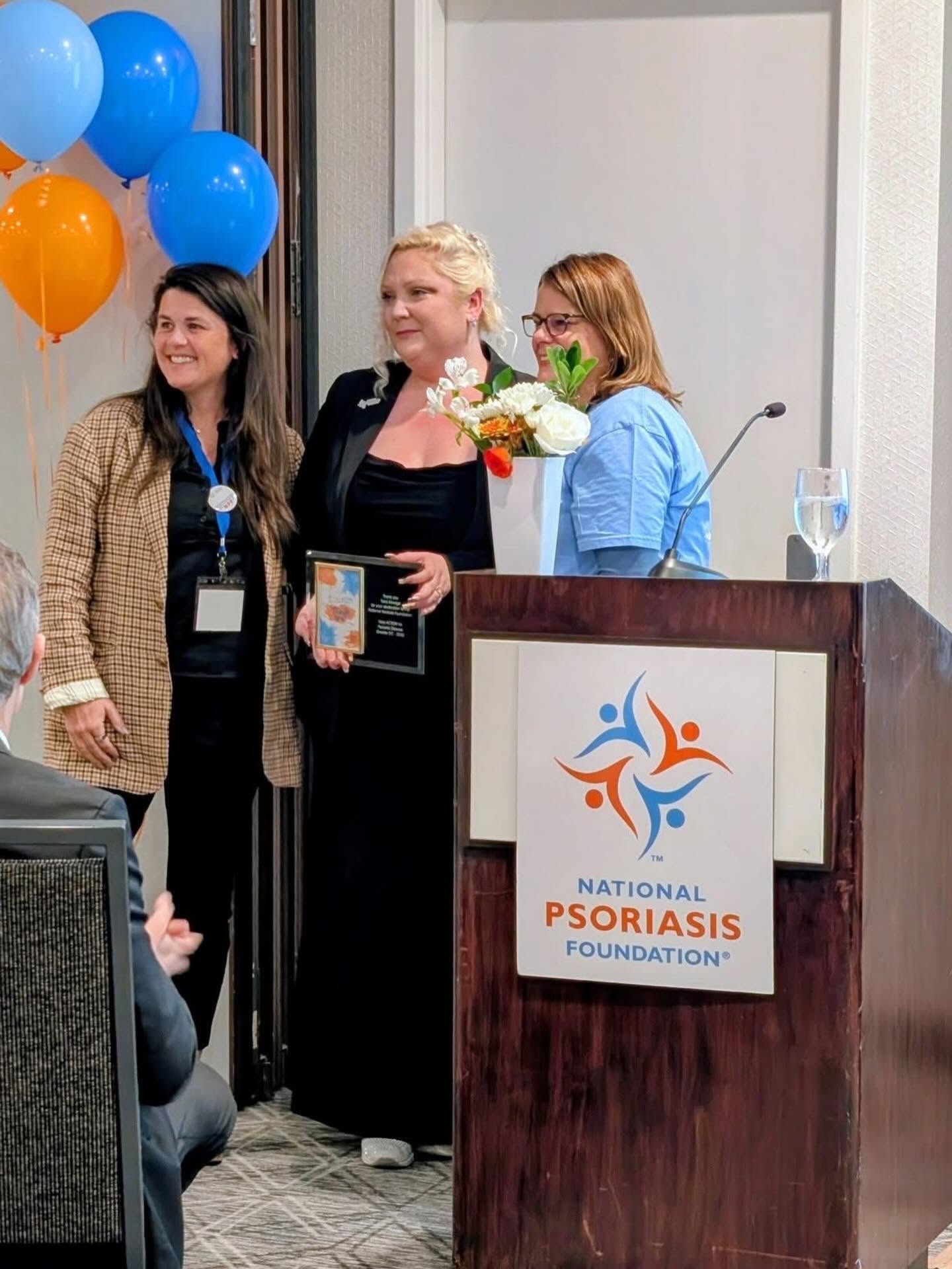 Three women standing together at a podium with balloons in the background, one holding a bouquet of flowers, smiling.