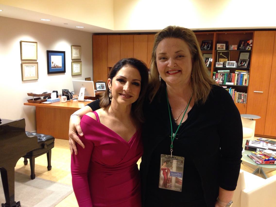 Two women standing close together, smiling, in an indoor setting with bookshelves and framed pictures in the background.