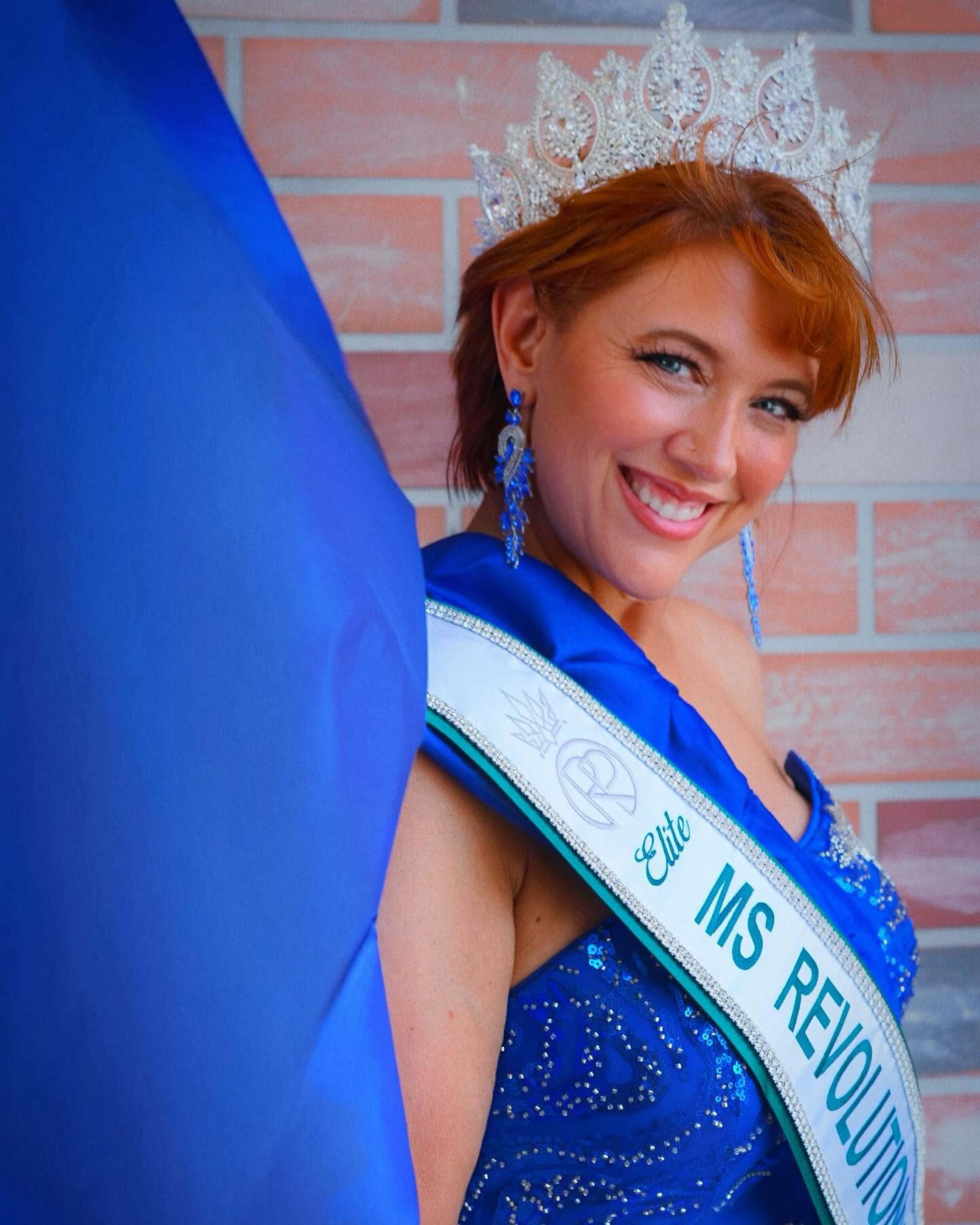 Young woman with red hair wearing a crown and sash, smiling, against a brick wall background.