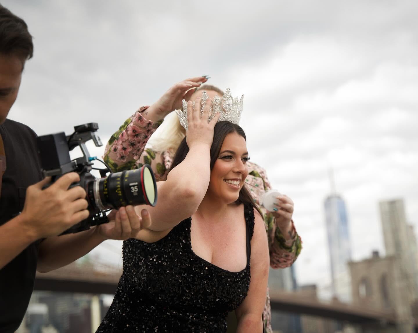 Woman smiling as a person places a crown on her head during an outdoor event, city skyline in background.