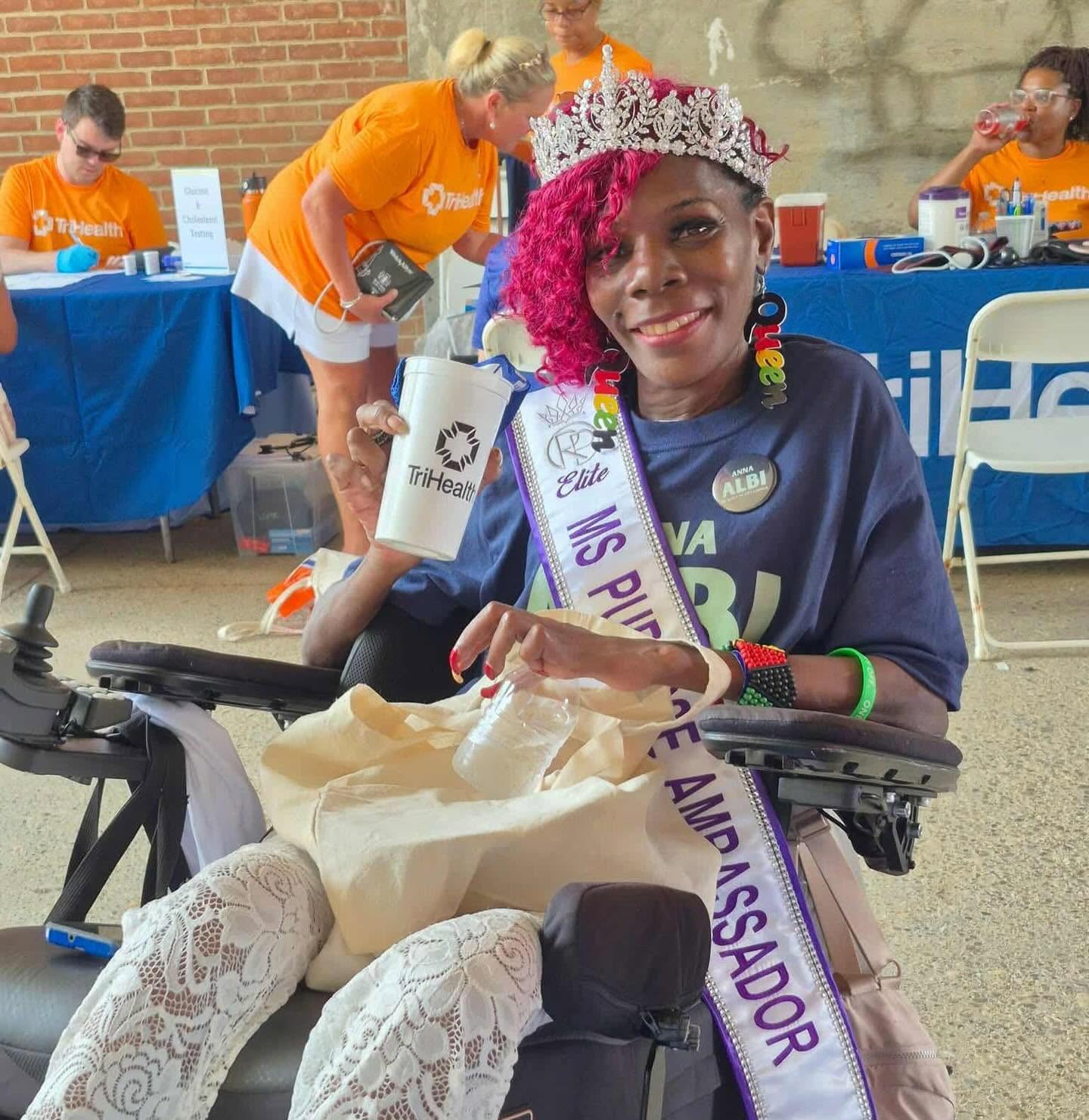 Young woman with pink hair wearing a crown and sash, smiling, sitting in a wheelchair outdoors, with people and tables in background.