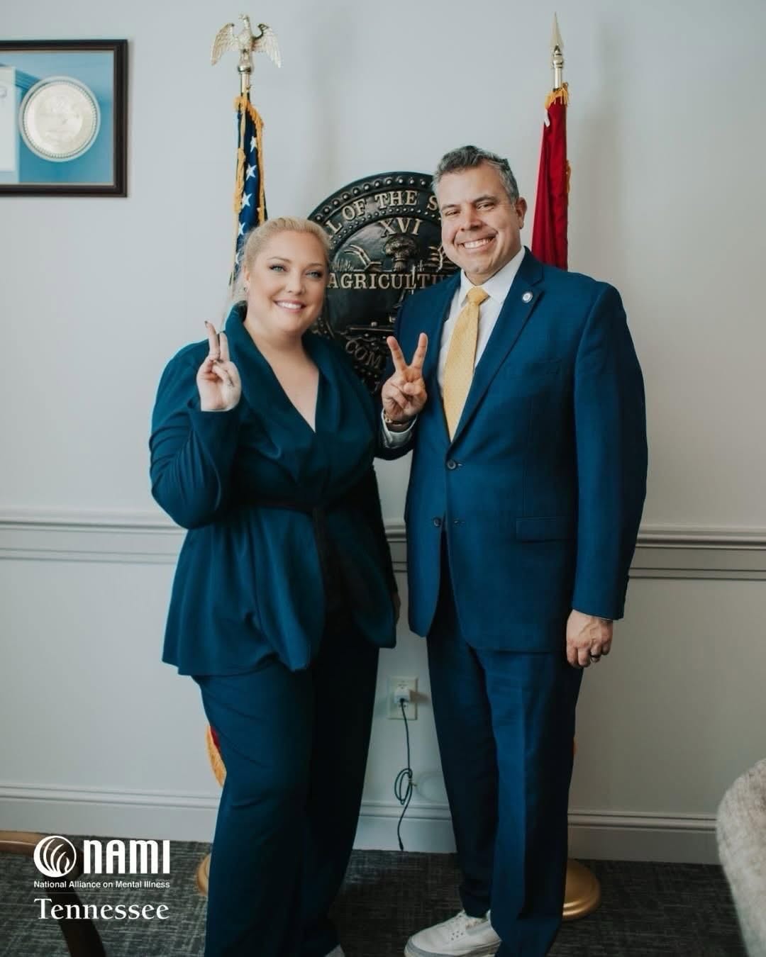 Two people in formal attire standing in front of a wall with flags and a seal, smiling and making peace signs.