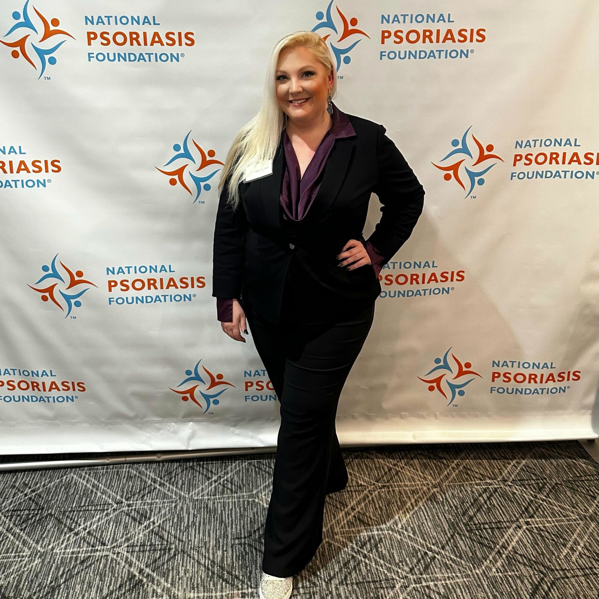 Woman in black suit standing in front of a backdrop with the National Psoriasis Foundation logo, smiling, with one hand on her hip.