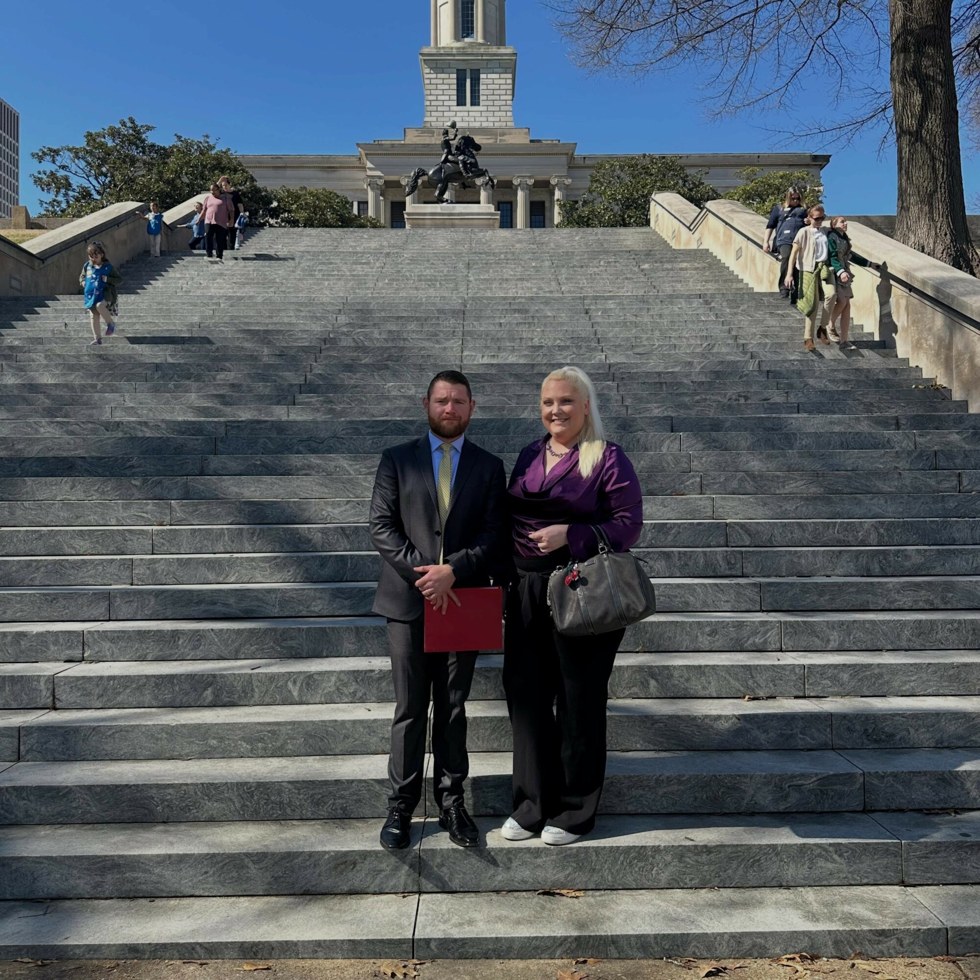 Two people, a man in a suit and a woman in a purple jacket, stand on stairs in front of a monument with a statue and people around. 20 words.