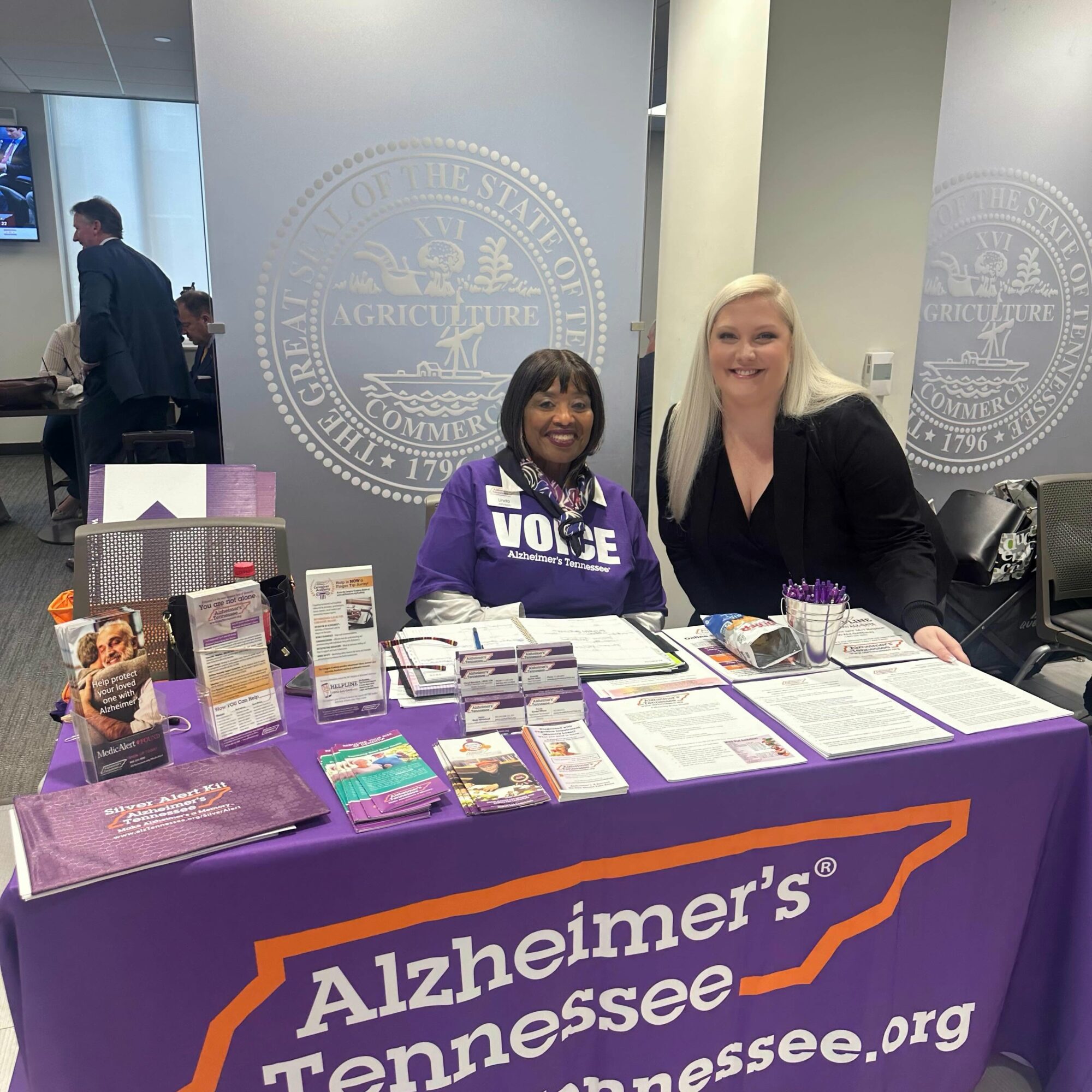 Two women sit behind a table with Alzheimer's Tennessee materials, smiling at the camera, in a conference room.