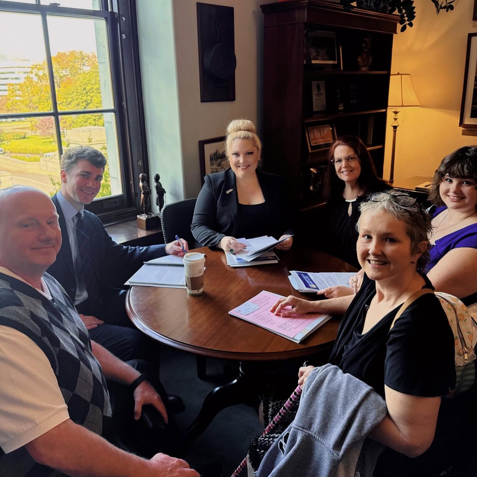 Six people sitting around a round table in a room with a window, bookshelves, and a lamp, smiling at the camera.